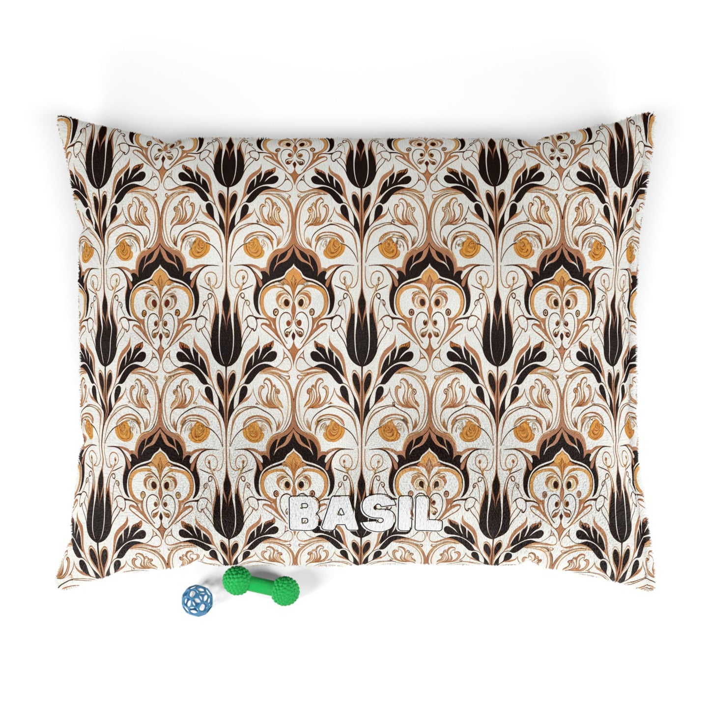 A rectangular pillow with an ornate African-inspired brown, gold, and black floral pattern. “BASIL” is embroidered in white letters at the bottom center. Two dog toys—a green bone and a blue ball—rest nearby on a white surface.