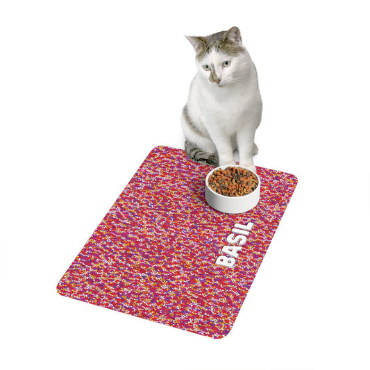 A white and gray cat sits on an African-inspired red, pink, and yellow patterned mat with a bowl of cat food. The mat has the name BASIL printed in white uppercase letters on the lower right side. The background is plain white.