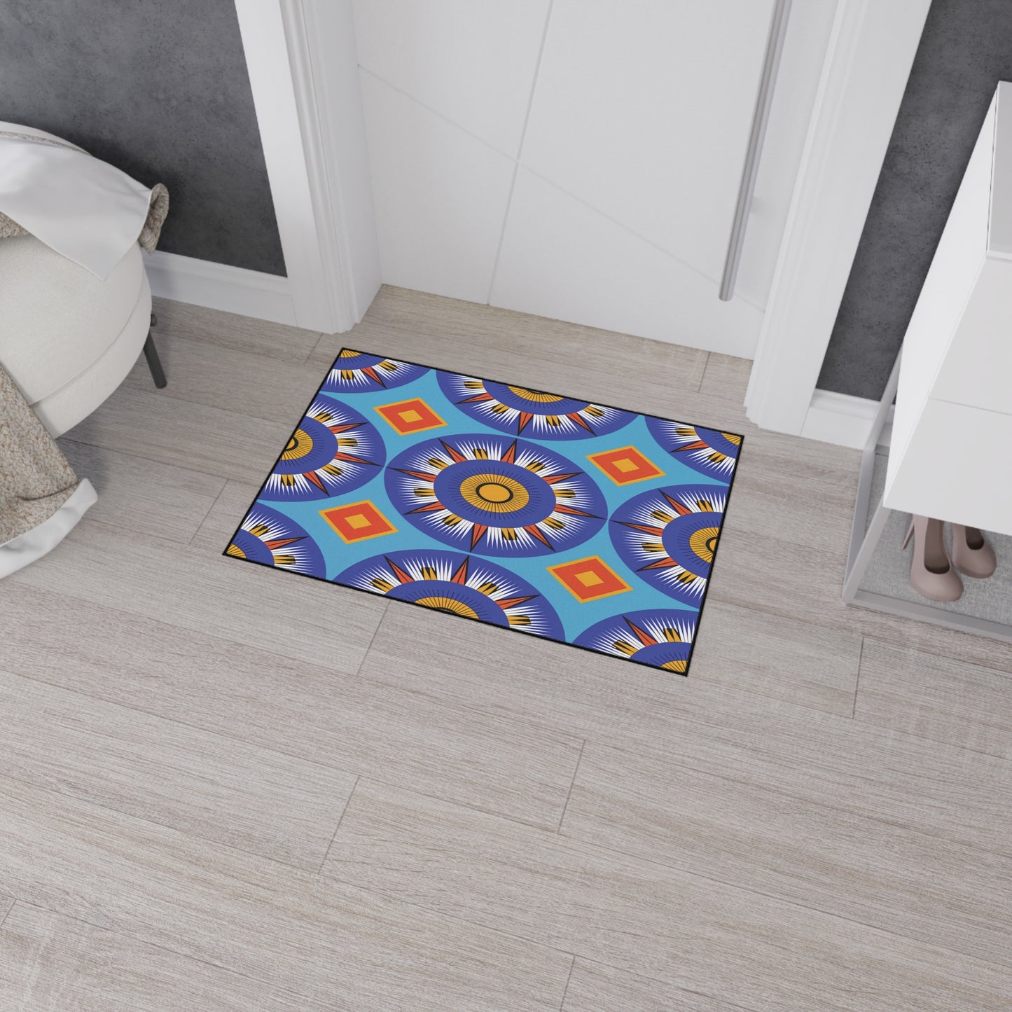 A colorful doormat with bold blue, orange, yellow, and red geometric sunburst patterns inspired by African-inspired decor lies on a light wood floor. It features non-slip rubber backing and sits near white walls, a shoe cabinet, and a bed with a white blanket.