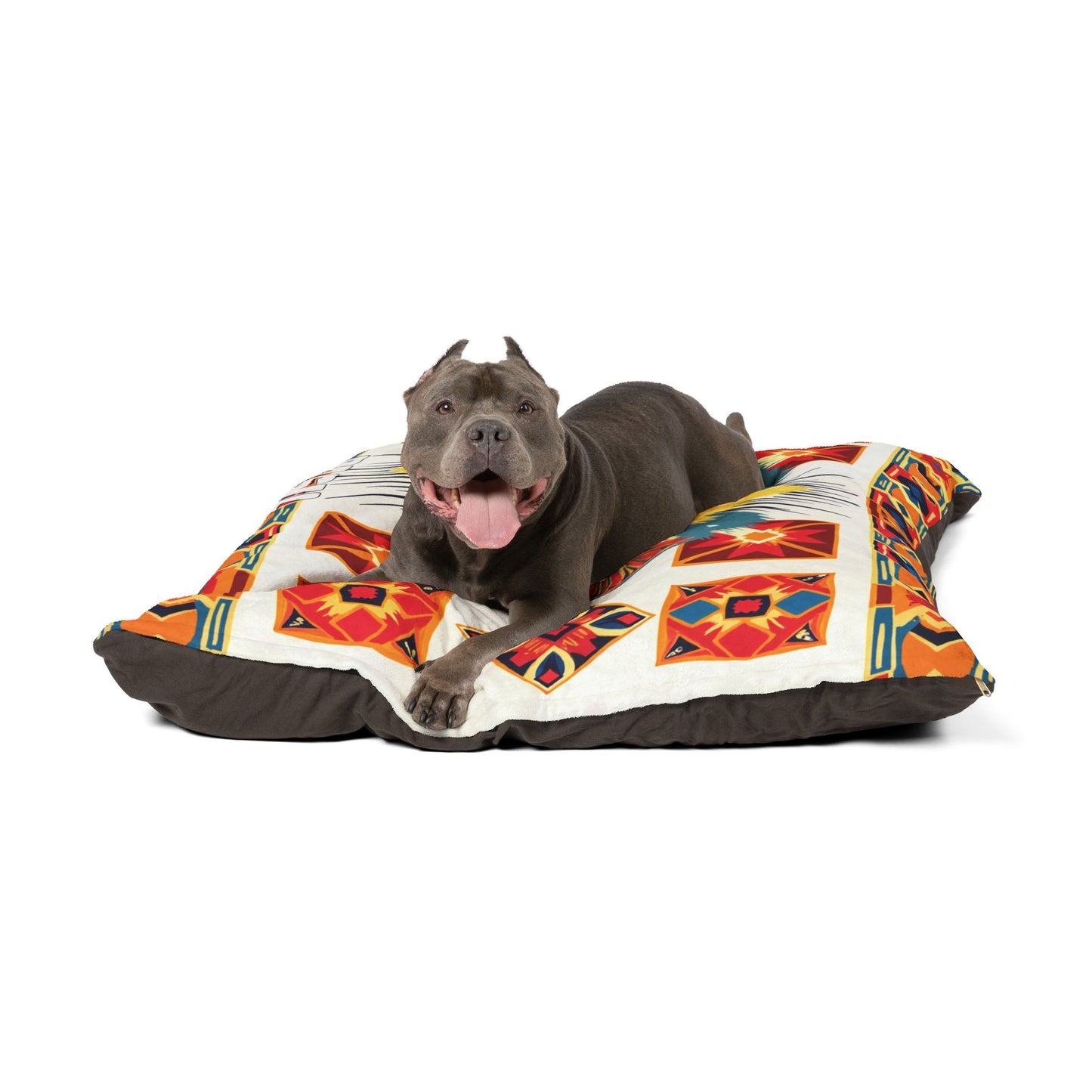 A happy gray pit bull dog with cropped ears lies on a large, African-inspired cushion, mouth open and tongue out, against a white background. The cushion features geometric orange, red, and yellow designs.