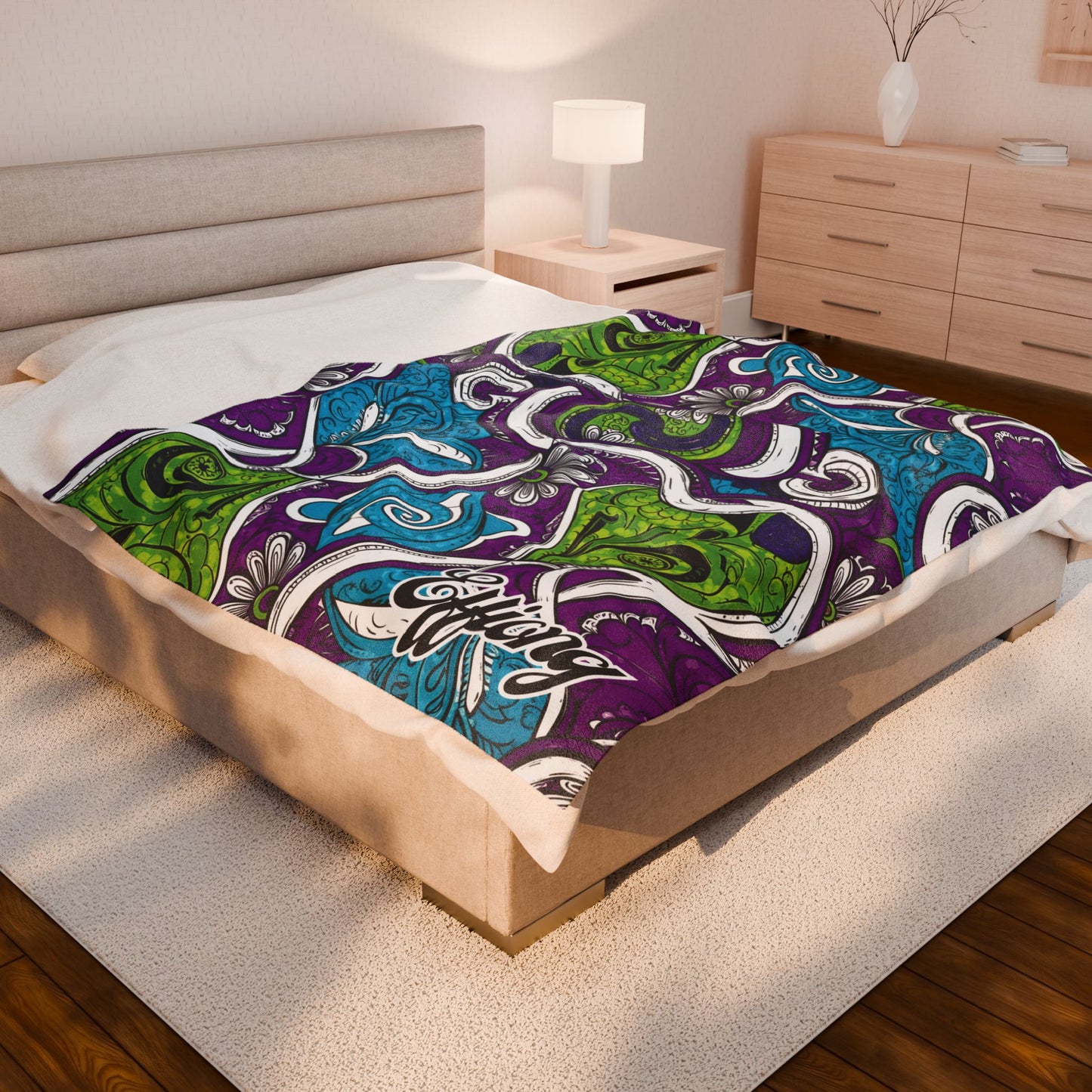 A modern bedroom features a large beige bed with an African-inspired blanket in blue, green, and purple. The room has wooden floors, a white rug, a nightstand with a lamp, and a dresser with a decorative vase. Warm lighting fills the space.