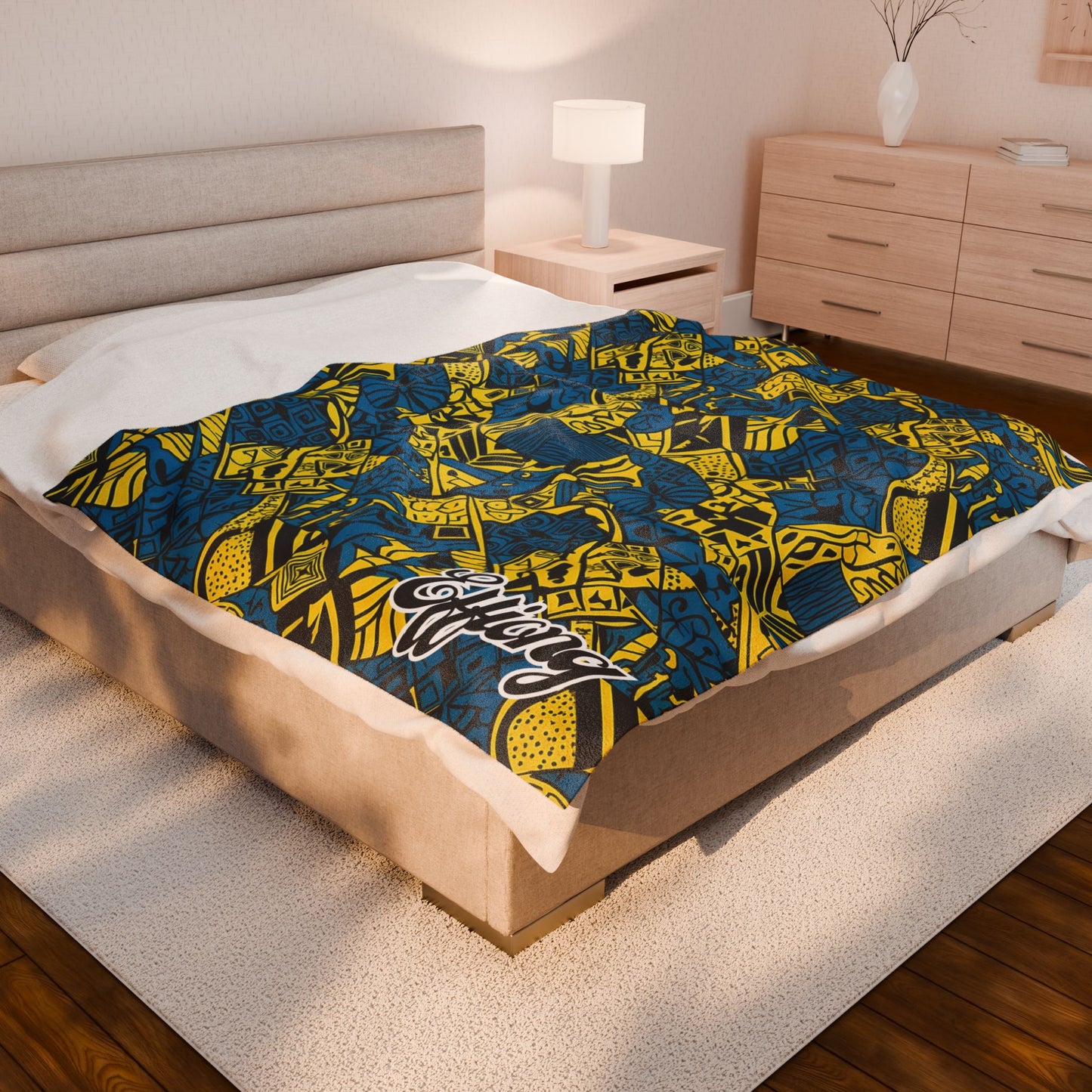 A modern bedroom featuring a beige upholstered bed with a vibrant, African-inspired blanket displaying abstract geometric shapes and “Melanin” in script on the corner. Warm wood floors, a nightstand, lamp, and dresser complete the look.