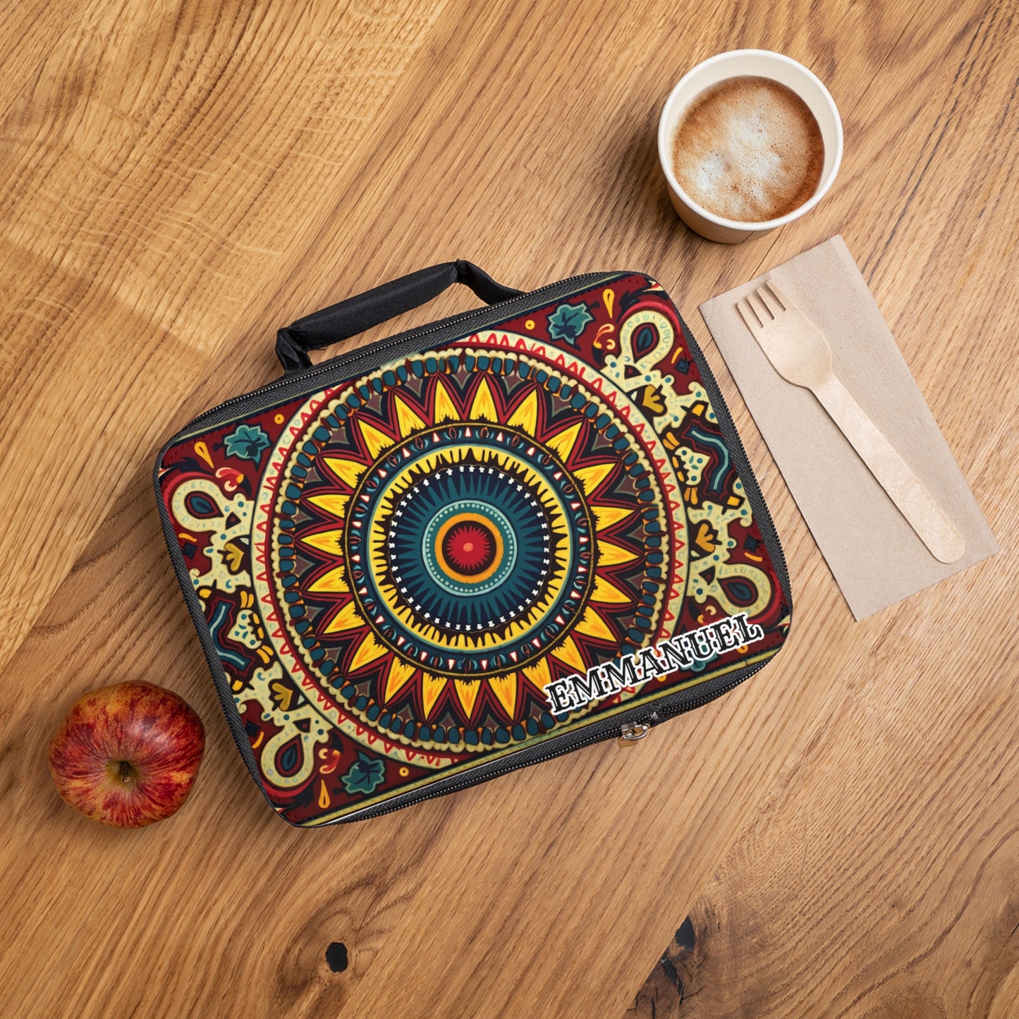 A vibrantly patterned African-inspired lunch bag with the name EMMANUEL sits on a wooden table, next to a red apple, a cardboard cup of frothy coffee, and a wooden fork and knife placed on a brown napkin.