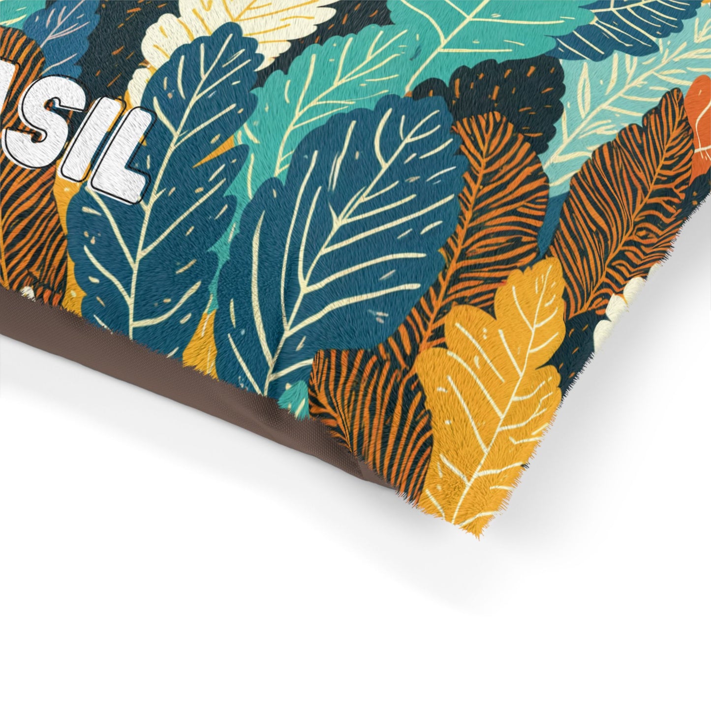 Close-up of a rectangular item, possibly a cushion or book, with a colorful leaf pattern in teal, green, yellow, and orange on fabric. Part of the word BASIL is visible on the left. The corner shows a brown edge and hints at durable construction.
