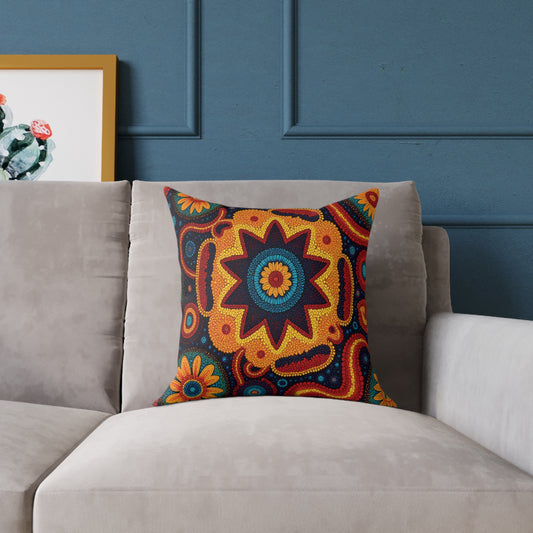A beige couch with a vibrant throw pillow featuring a bold, symmetrical floral and geometric design in red, yellow, orange, and blue. Behind the couch is a blue wall with molding and part of a framed cactus picture is visible on the left.