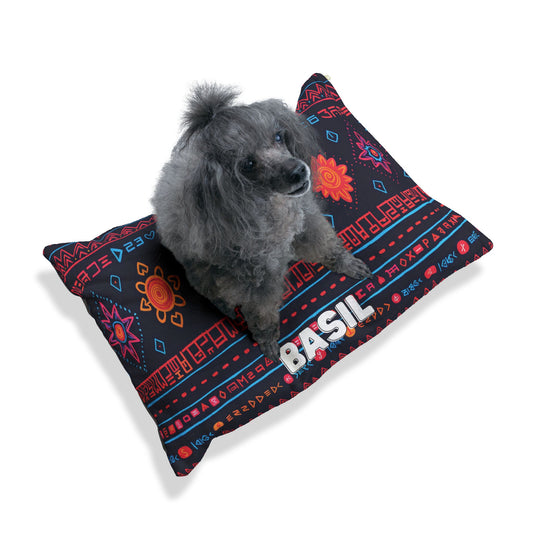A small gray poodle with curly fur sits on a personalized pet bed featuring bright geometric and floral patterns. The durable construction of the mostly black pillow includes BASIL in large white letters along the front edge.