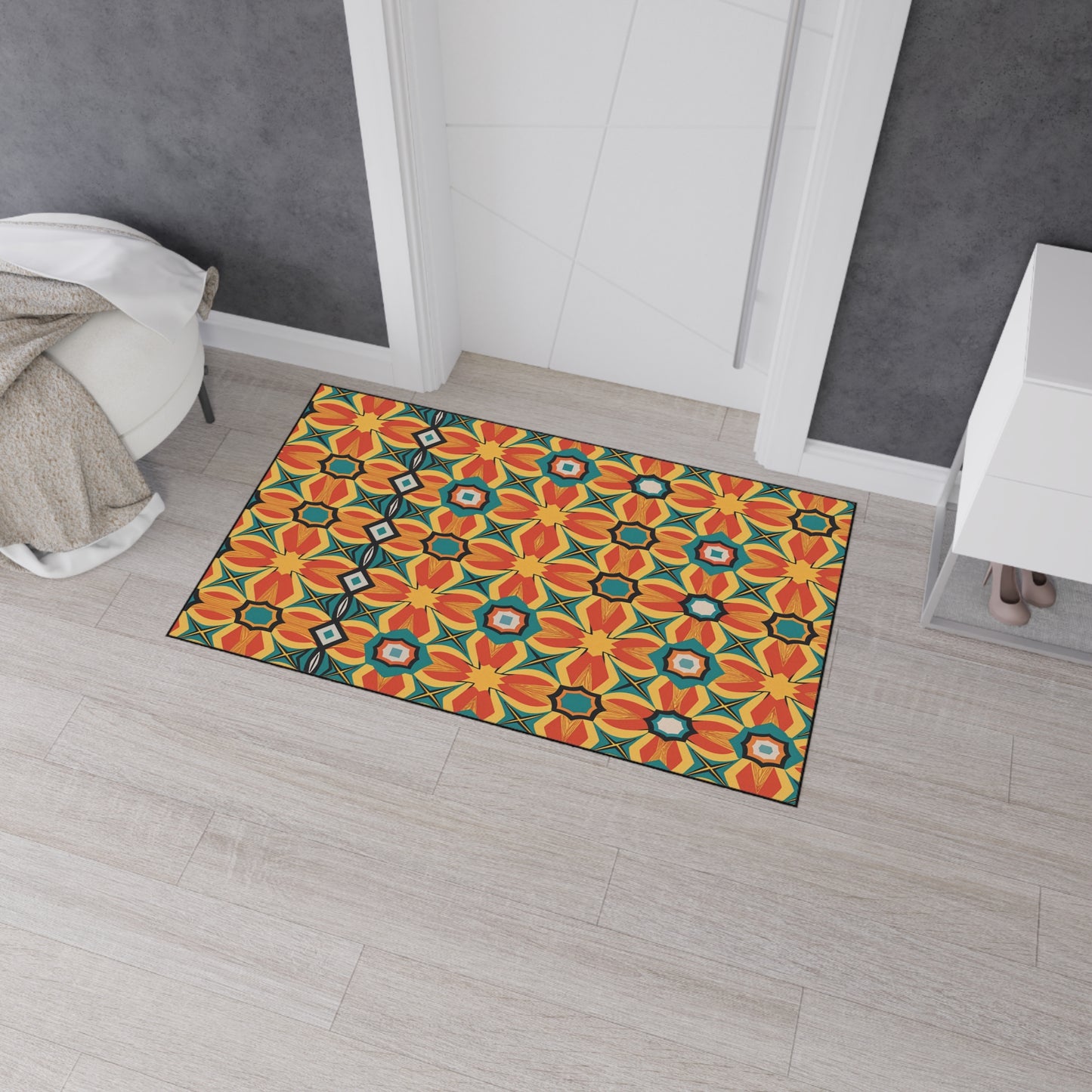 A rectangular mat with African-inspired aesthetics and a colorful geometric floral pattern in orange, yellow, black, and teal lies on a light wood floor near a white door. A beige throw blanket and a white cabinet complete the modern entryway.