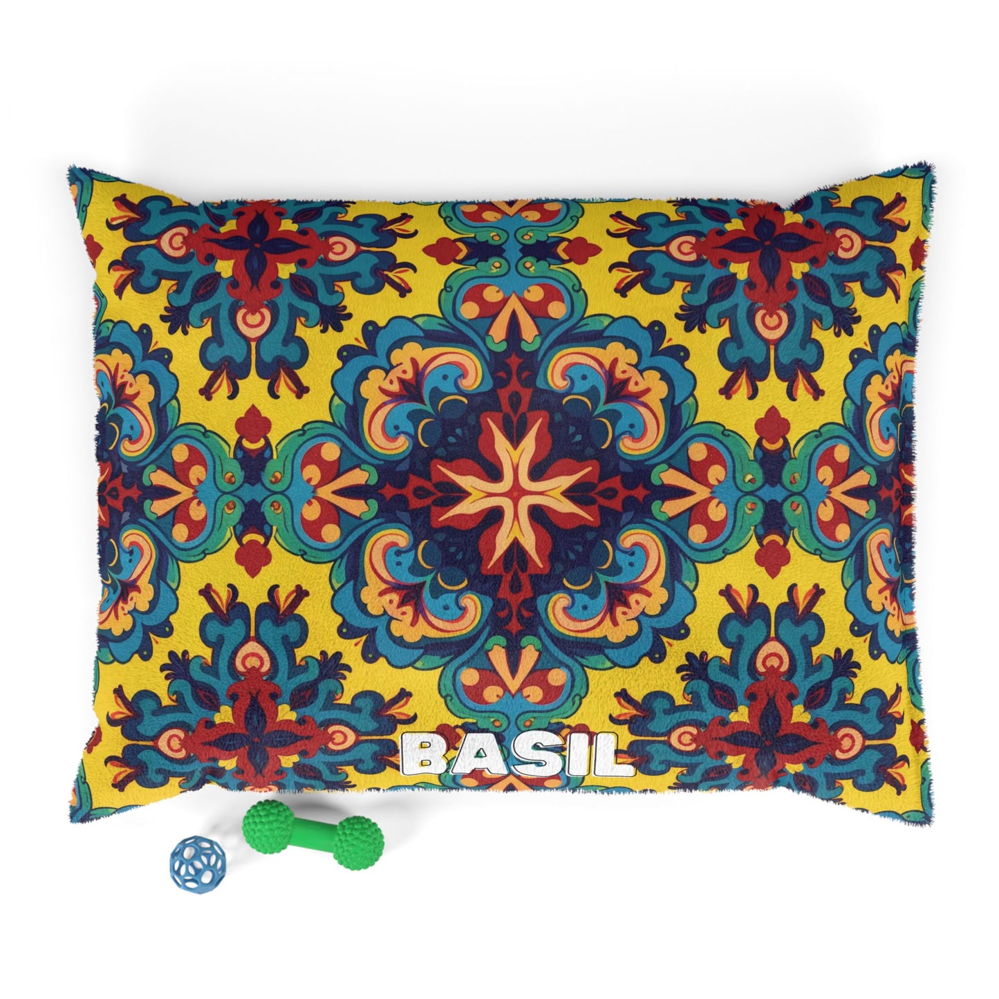 A cozy pet pillow featuring a bold, colorful floral and geometric pattern in yellow, red, teal, and navy. “BASIL” is spelled in white at the bottom. Nearby are two dog toys: a green bone and a small blue ball.