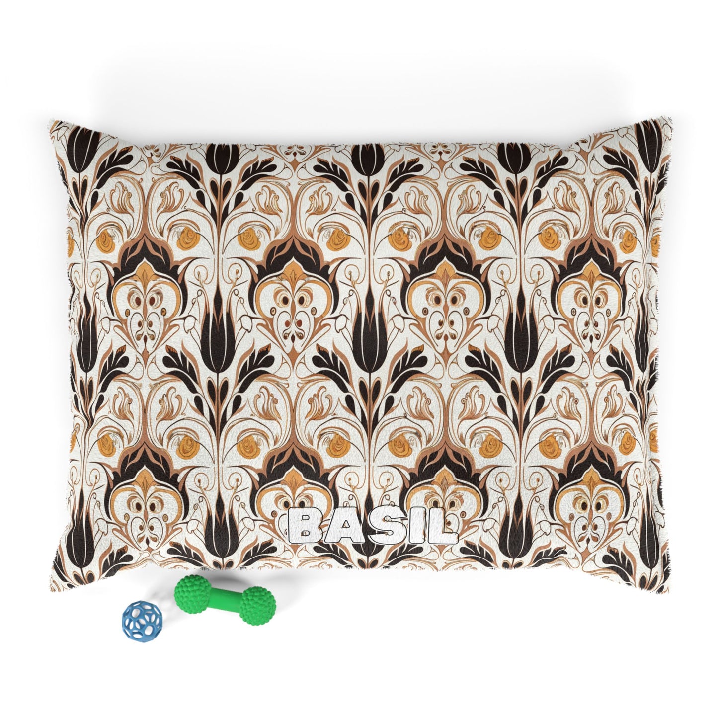 A rectangular pillow with an African-inspired, art nouveau-style floral pattern in brown, gold, and black on a cream background. The name “BASIL” is printed in white at the bottom. Two dog toys—a blue ball and a green bone—are placed below.