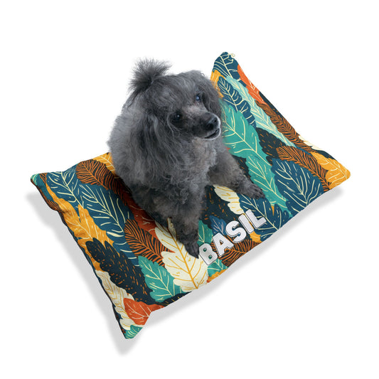 A small, curly-haired gray poodle sits on a colorful pillow with a leafy pattern in green, yellow, orange, and black. The word BASIL is printed in bold white letters on this personalized pet bed, crafted for luxurious comfort. The background is plain white.