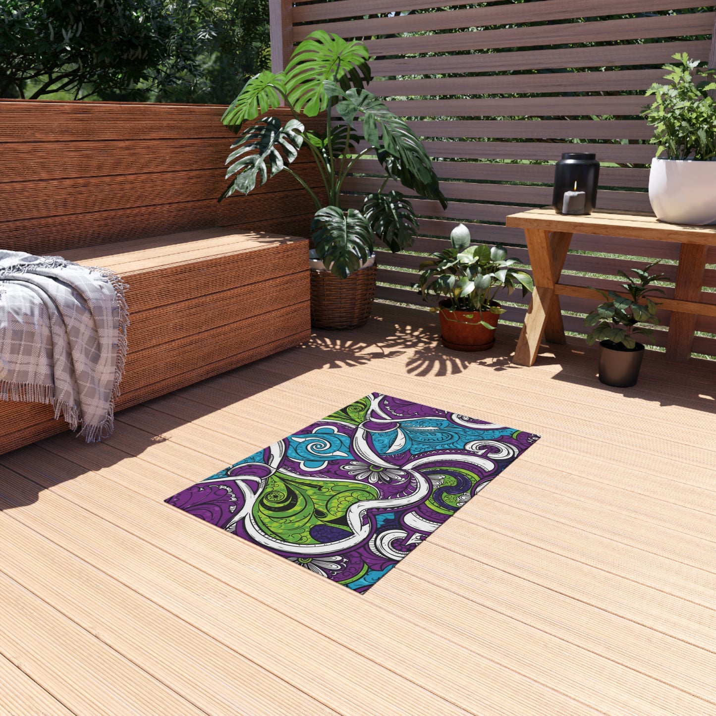 Outdoor patio with wooden flooring, a wicker bench with a gray plaid blanket, potted green plants, and a small wooden table topped with a black pitcher. An African-inspired outdoor rug featuring purple, green, and blue swirls adds vibrant flair to the space.