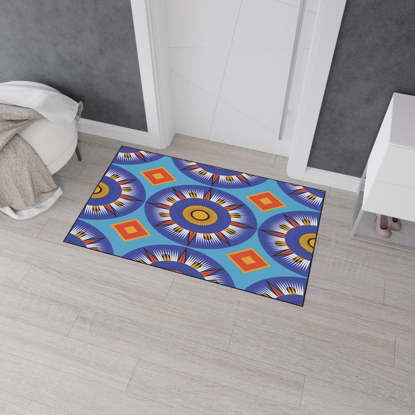 A colorful, African-inspired doormat with bold, circular geometric patterns in blue, orange, yellow, and white lies on a light wood floor near a white door. Gray walls, a white cabinet, and a beige textured throw complete the scene.