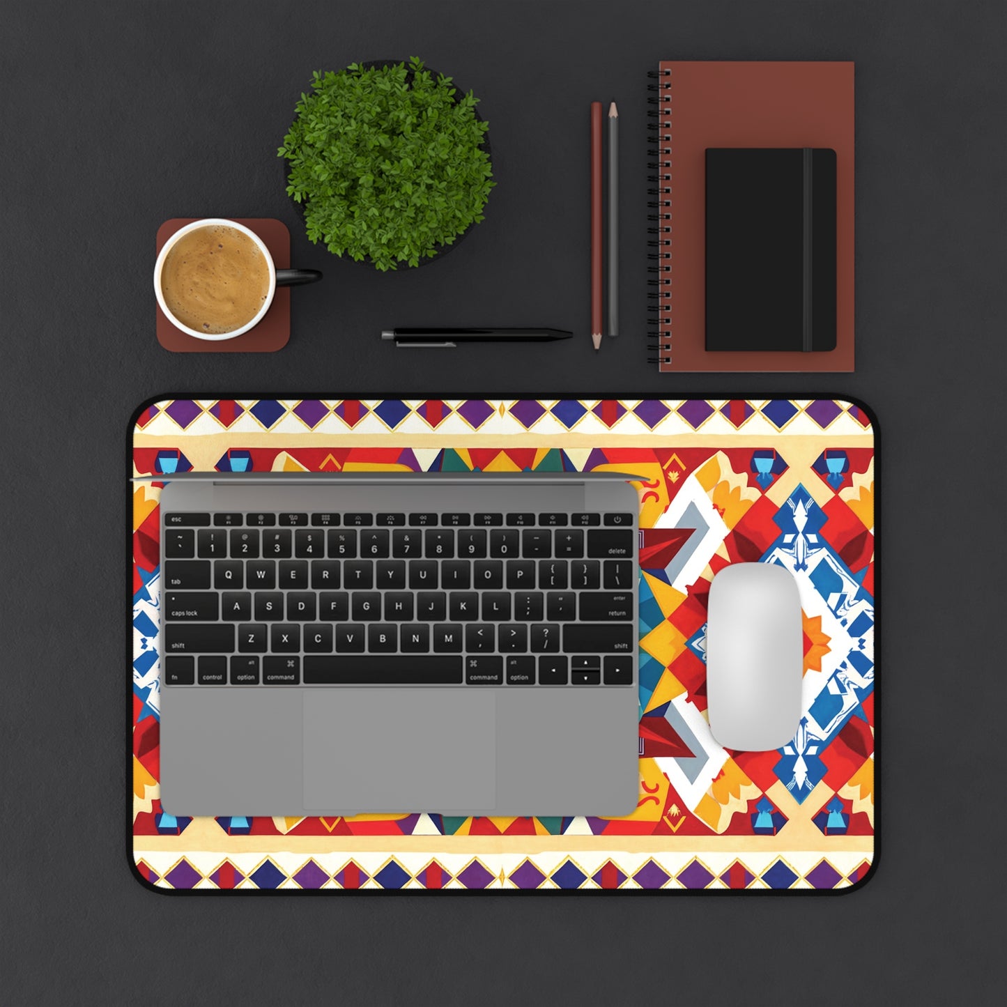 A laptop and white mouse sit on a colorful geometric-patterned Neoprene desk mat atop a dark desk. Nearby are a cup of coffee, a potted plant, writing tools, and two notebooks—one brown, one black—arranged neatly above the mat.