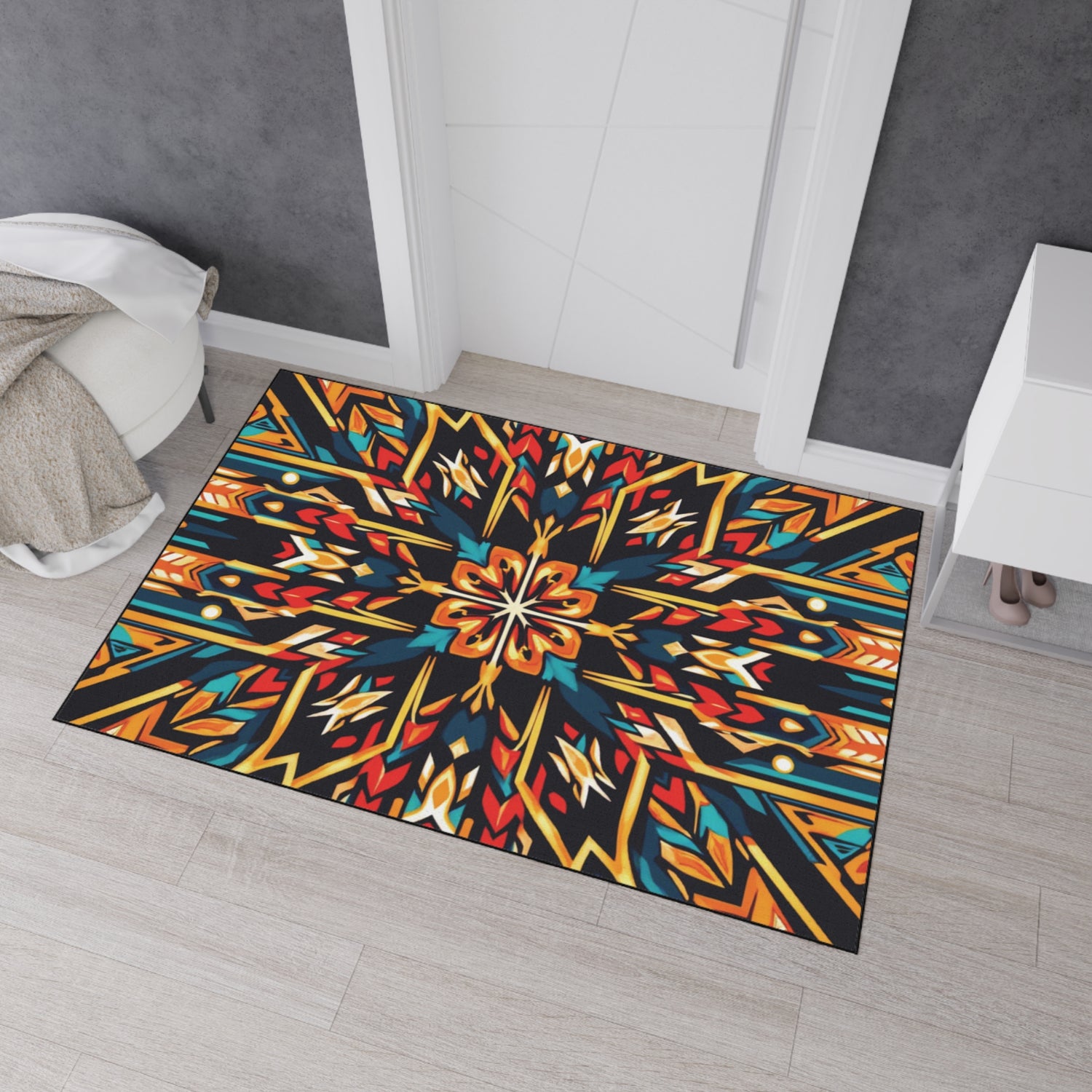 A rectangular, African-inspired rug featuring a vibrant, symmetrical geometric pattern with red, yellow, blue, and orange hues lies on a light wood floor near a doorway. The rug is surrounded by gray and white walls and a white cabinet.
