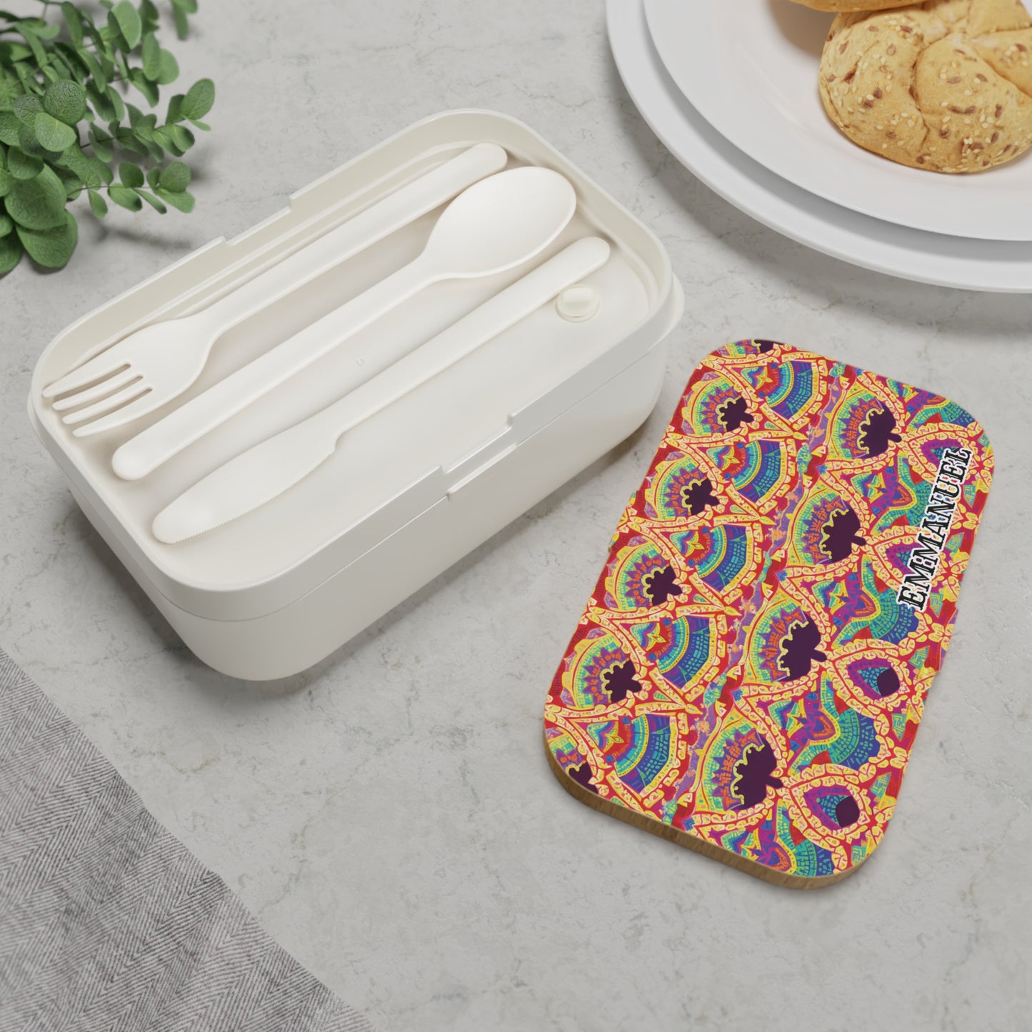A white, BPA-free lunchbox with a fork, spoon, and knife rests on a light marble surface. Its colorful lid features a vibrant, abstract geometric pattern inspired by African art and the words “EAT ANYWHERE.” Nearby are a plant and cookies.