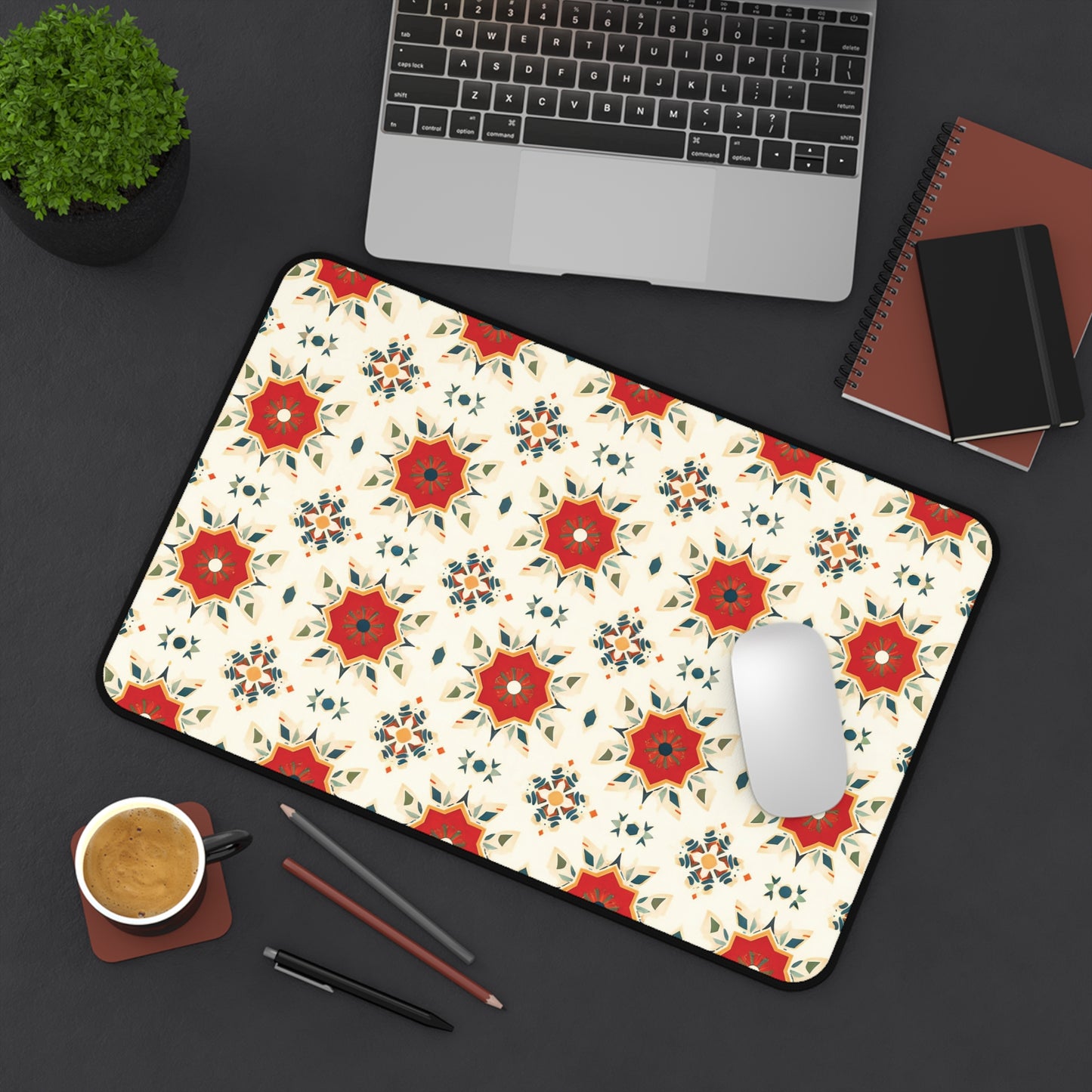 A large Neoprene desk mat with a red and orange geometric floral pattern on a cream background brightens up the dark desk—a colorful workspace accessory that holds a laptop, notebooks, smartphone, potted plant, coffee cup, pencils, and mouse.