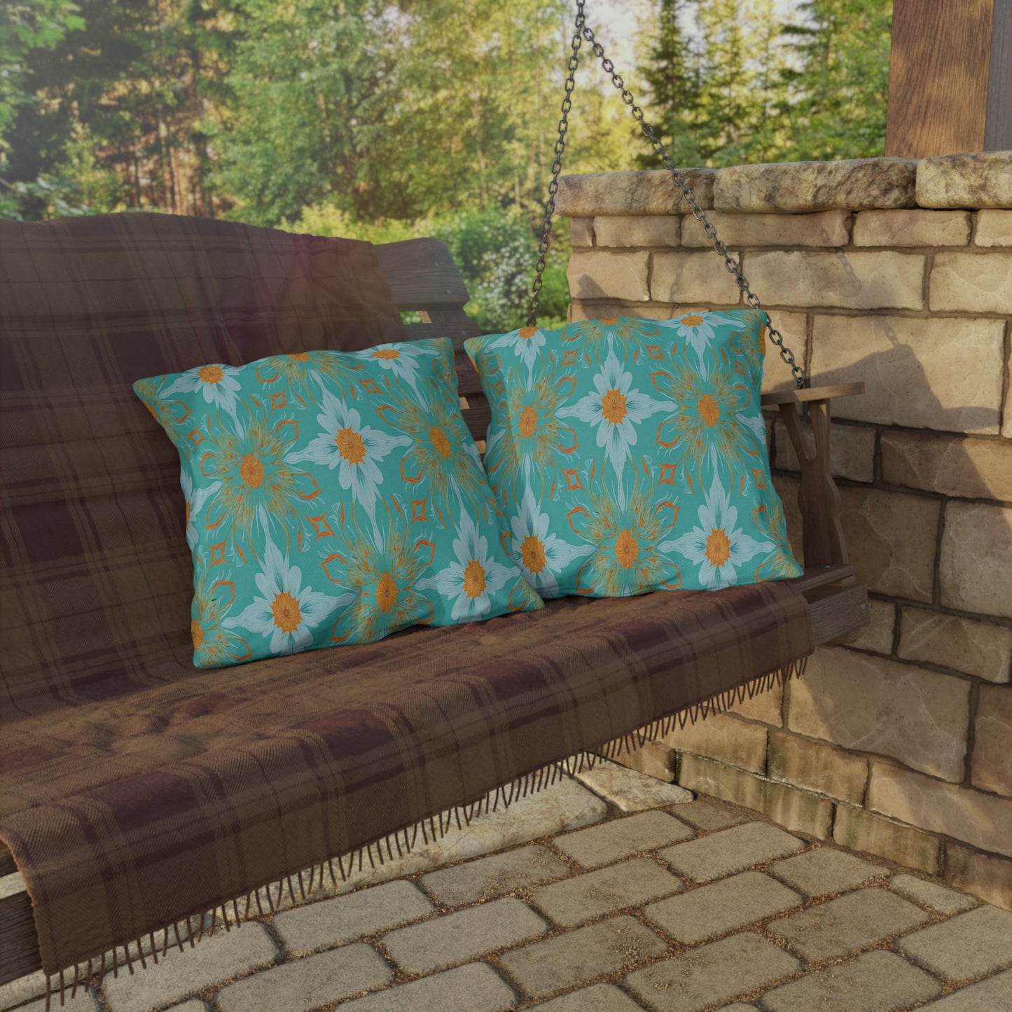 A wooden porch swing with a brown plaid blanket draped over it holds two teal cushions featuring African-inspired orange and white floral patterns. The swing hangs by chains near a stone wall on a patio, with green trees in the background.