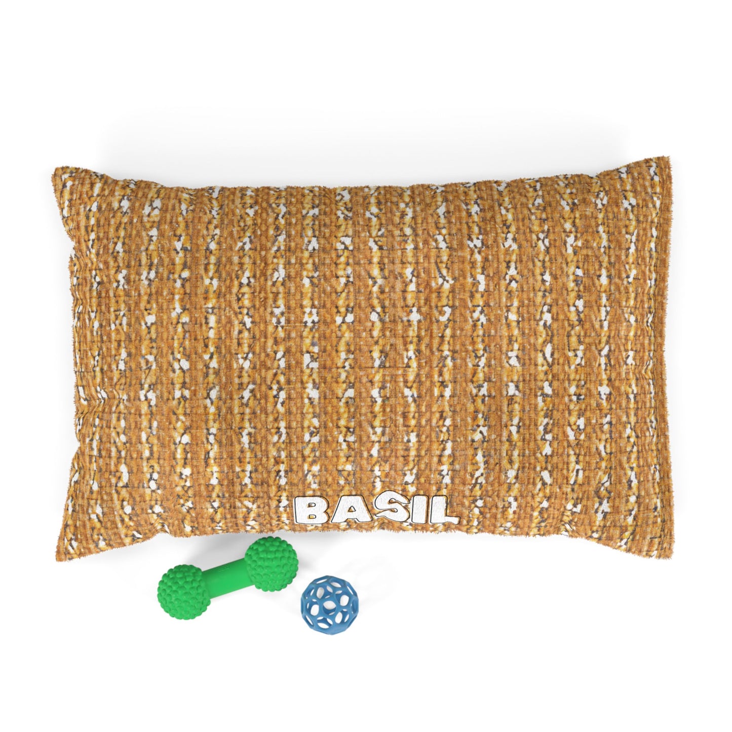 A brown woven custom dog bed labeled BASIL in white letters sits atop a white surface. In front of the bed are two dog toys: a green dumbbell-shaped toy and a blue ball with open lattice-like holes.