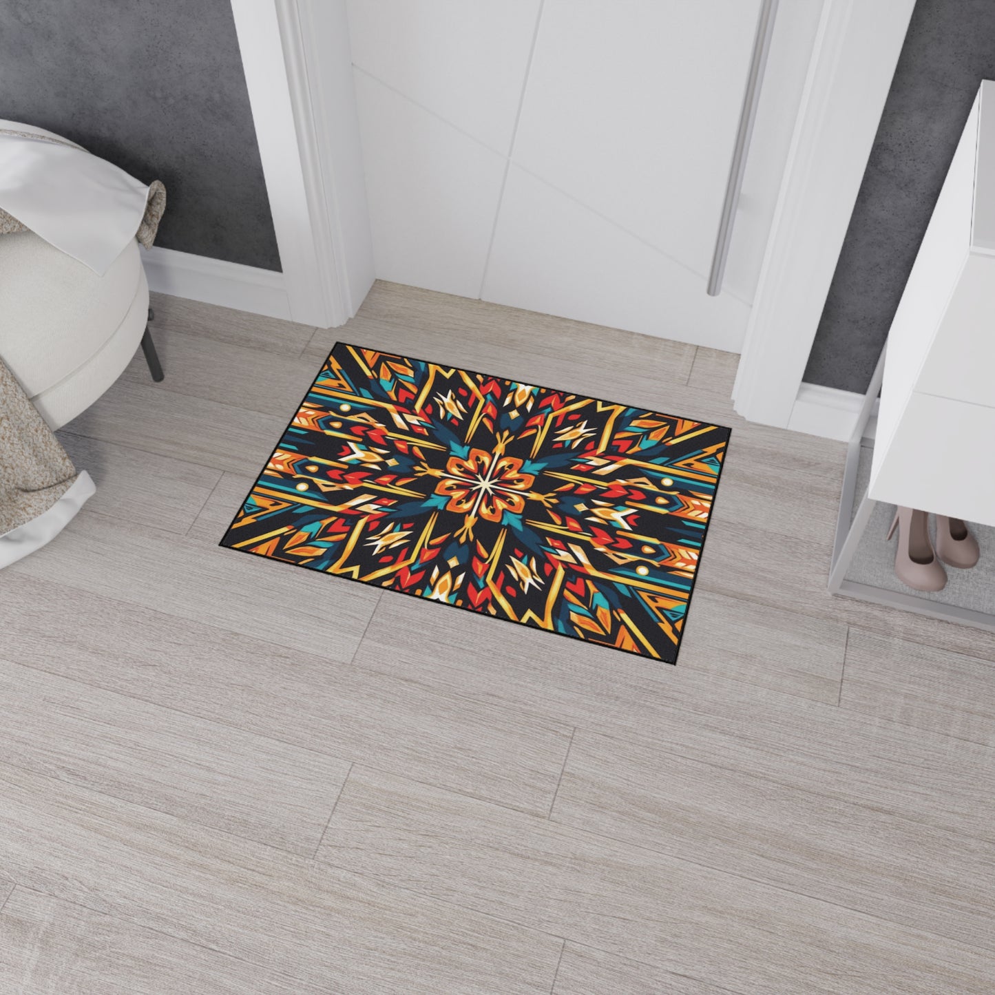 A colorful, African-inspired doormat with a symmetrical, kaleidoscopic geometric pattern in blue, orange, yellow, and red lies on light wood flooring near a white door. The room has a white bench, gray wall, and cabinet with visible shoes inside.