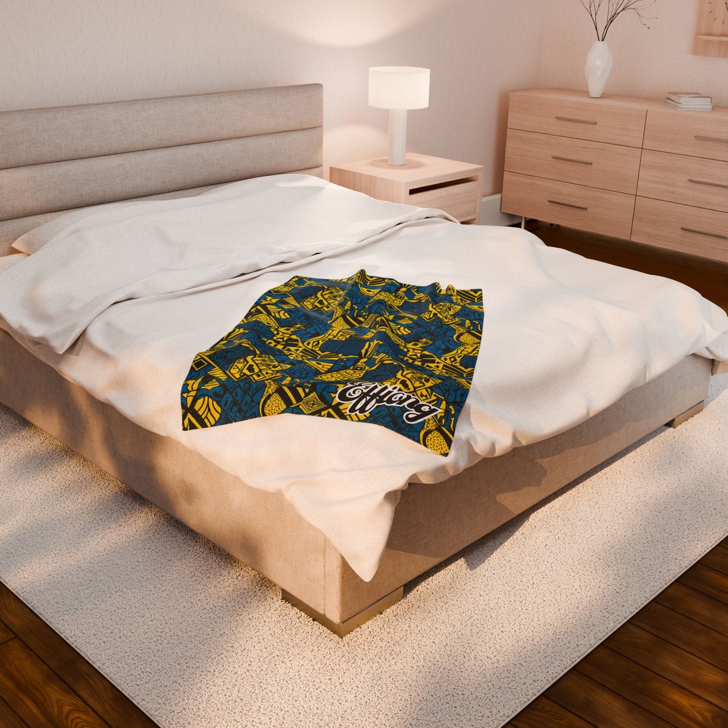 A modern bedroom with a beige upholstered bed, white bedding, and an African-inspired blanket lying on the blanket. The room features wooden floors, a dresser, a nightstand with a lamp, and a vase with branches.