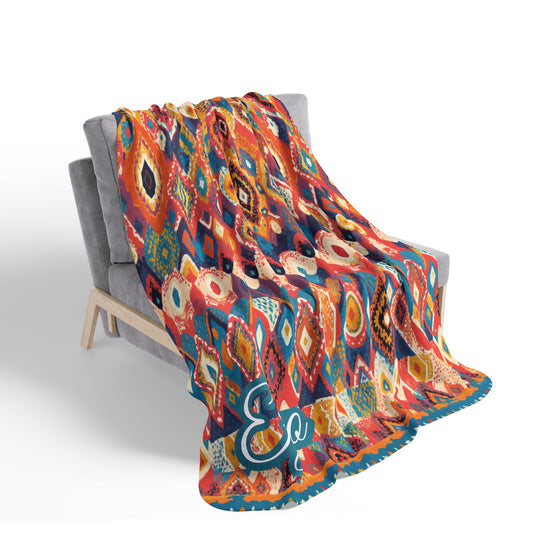 A gray cushioned chair holds a vibrant, African-inspired blanket with a bold, multicolored geometric pattern in red, blue, yellow, and orange. The lower edge of the blanket features a stylized turquoise script, possibly the letters Er.