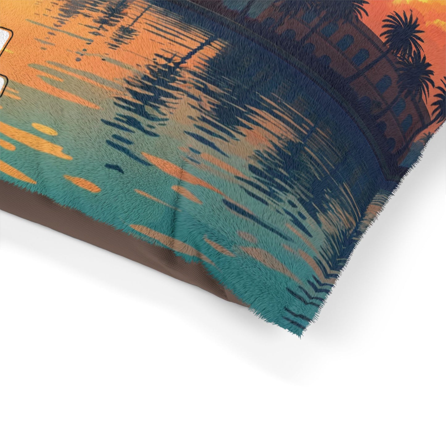 A close-up of a fabric item—possibly a personalized pet bed—featuring a vibrant sunset scene with palm trees, buildings, and reflective water in orange, teal, and blue hues. The fabric shows slightly fuzzy edges for an added touch of luxurious comfort.