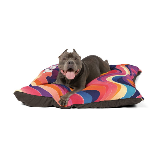 A gray dog with cropped ears lies on a large, colorful cushion with a swirling rainbow pattern, enjoying luxurious comfort. The happy pup stretches its front paws forward on the bed, which boasts durable construction against a plain white background.