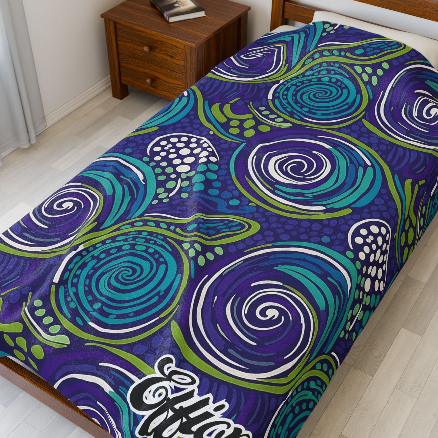 A single bed features a colorful African-inspired blanket with swirling abstract patterns in blue, teal, green, and white. The bed sits beside a wooden nightstand with books, in a bright room with light flooring and white curtains.