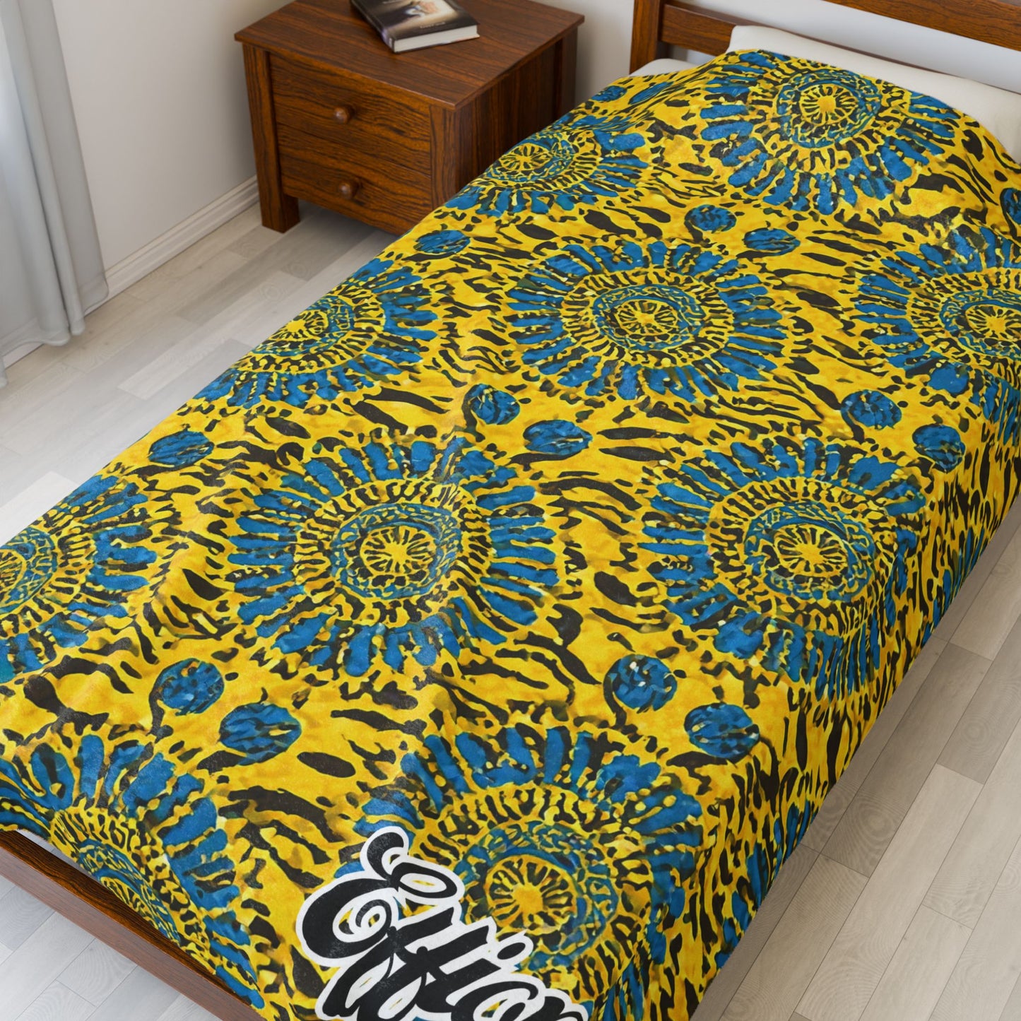A single bed with a bright yellow and blue African-inspired blanket featuring circular, sunburst-like designs and animal print motifs. The word “Effort” is visible at the bottom corner. A wooden nightstand beside the bed holds books.