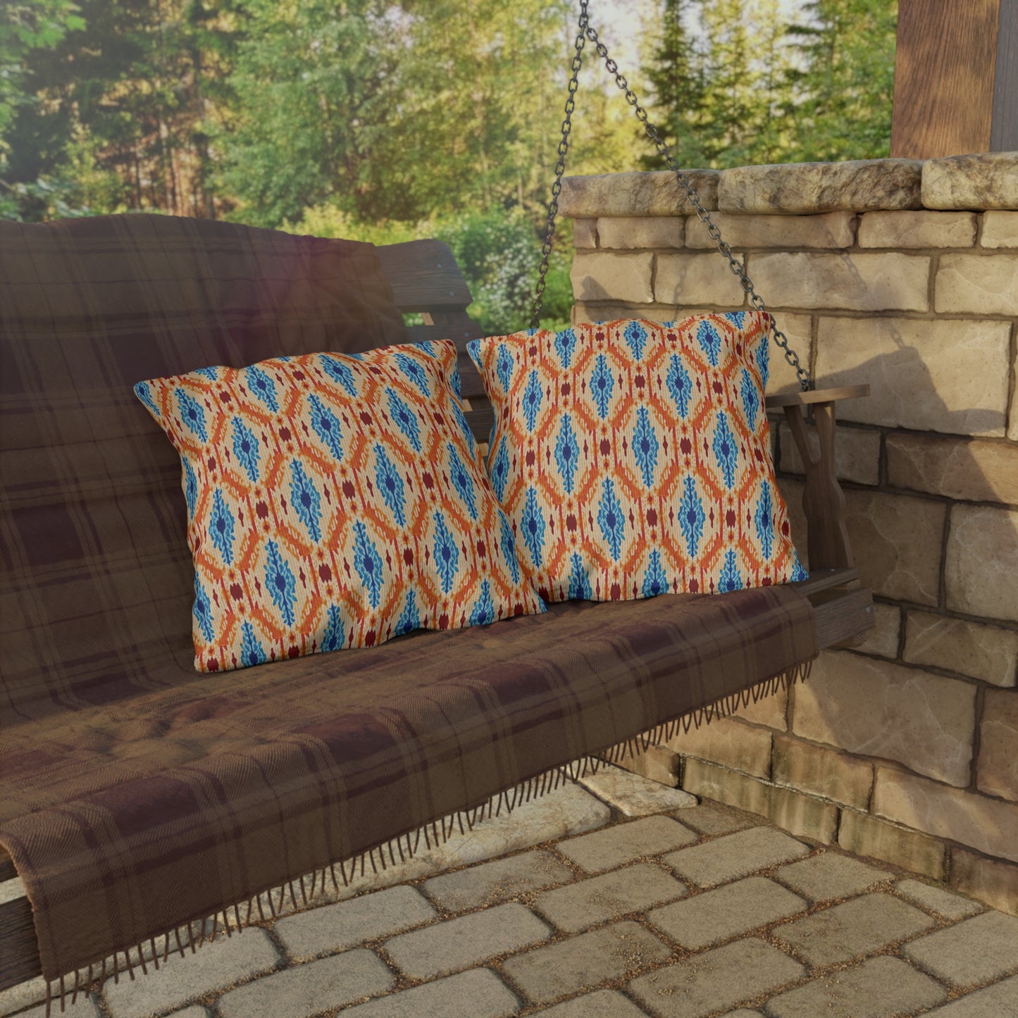 A wooden porch swing with a plaid blanket and two colorful outdoor pillows sits on a stone patio near a stacked stone wall, with trees and greenery visible in the background.