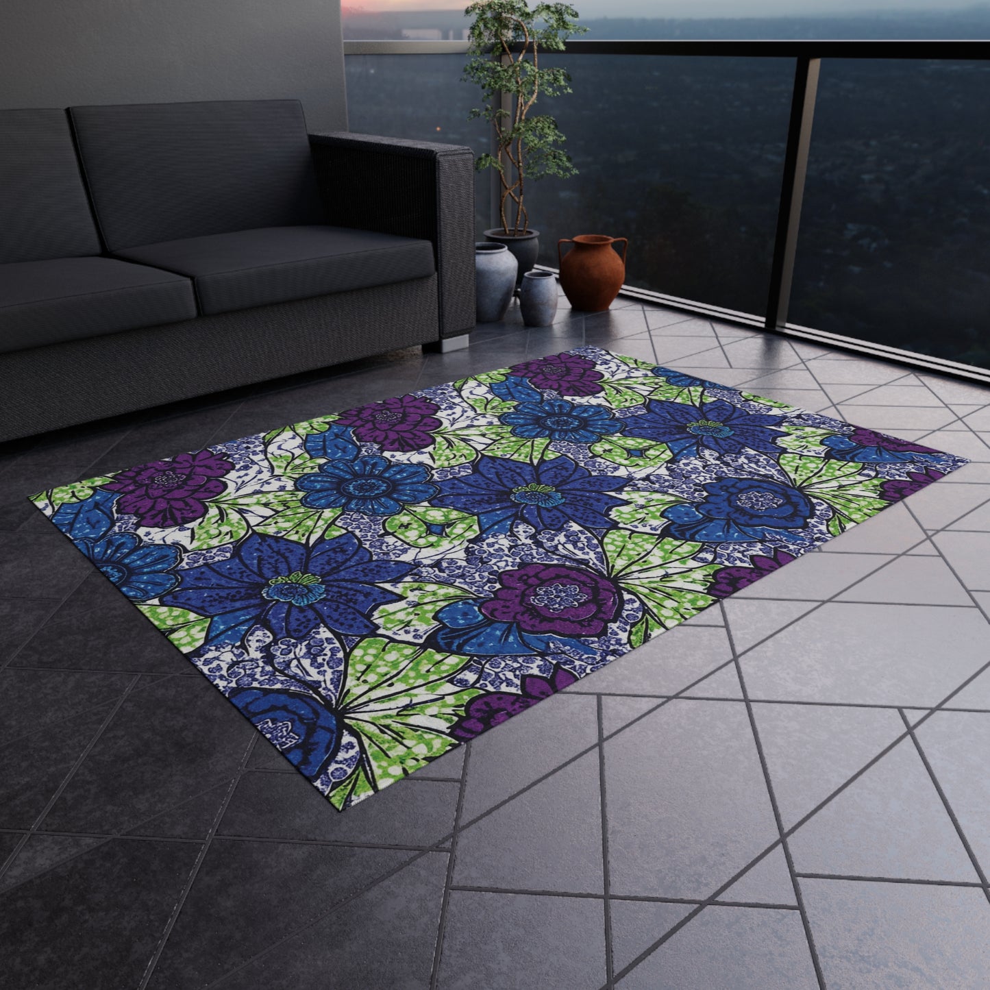 A colorful rectangular polyester chenille rug with bold floral patterns in blue, purple, green, and white sits on a tiled balcony floor. A gray sofa, potted plants, and a cityscape view through glass railing are in the background at dusk.