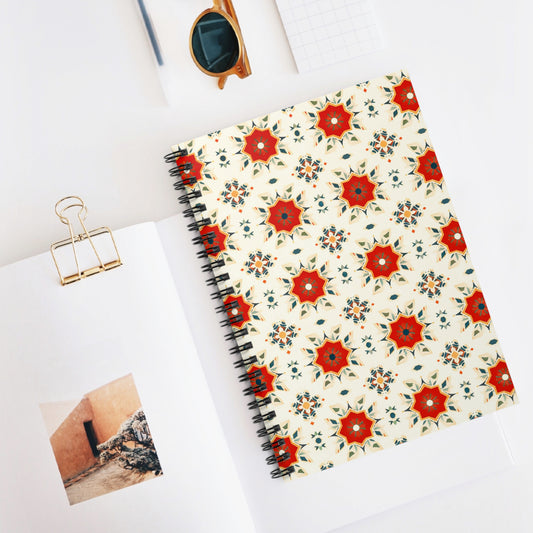 A premium African-inspired spiral notebook with a decorative red and blue geometric star pattern lies on a white desk beside an open book, sunglasses, a gold paper clip, and a small grid-patterned notepad.
