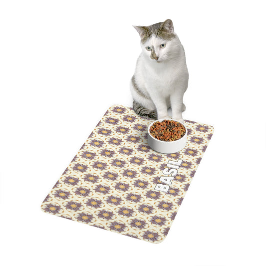 A white and gray cat sits next to a bowl of dry cat food on an African-inspired patterned mat. The mat features cat faces and the name BASIL printed in large white letters on one side. The background is plain and white.