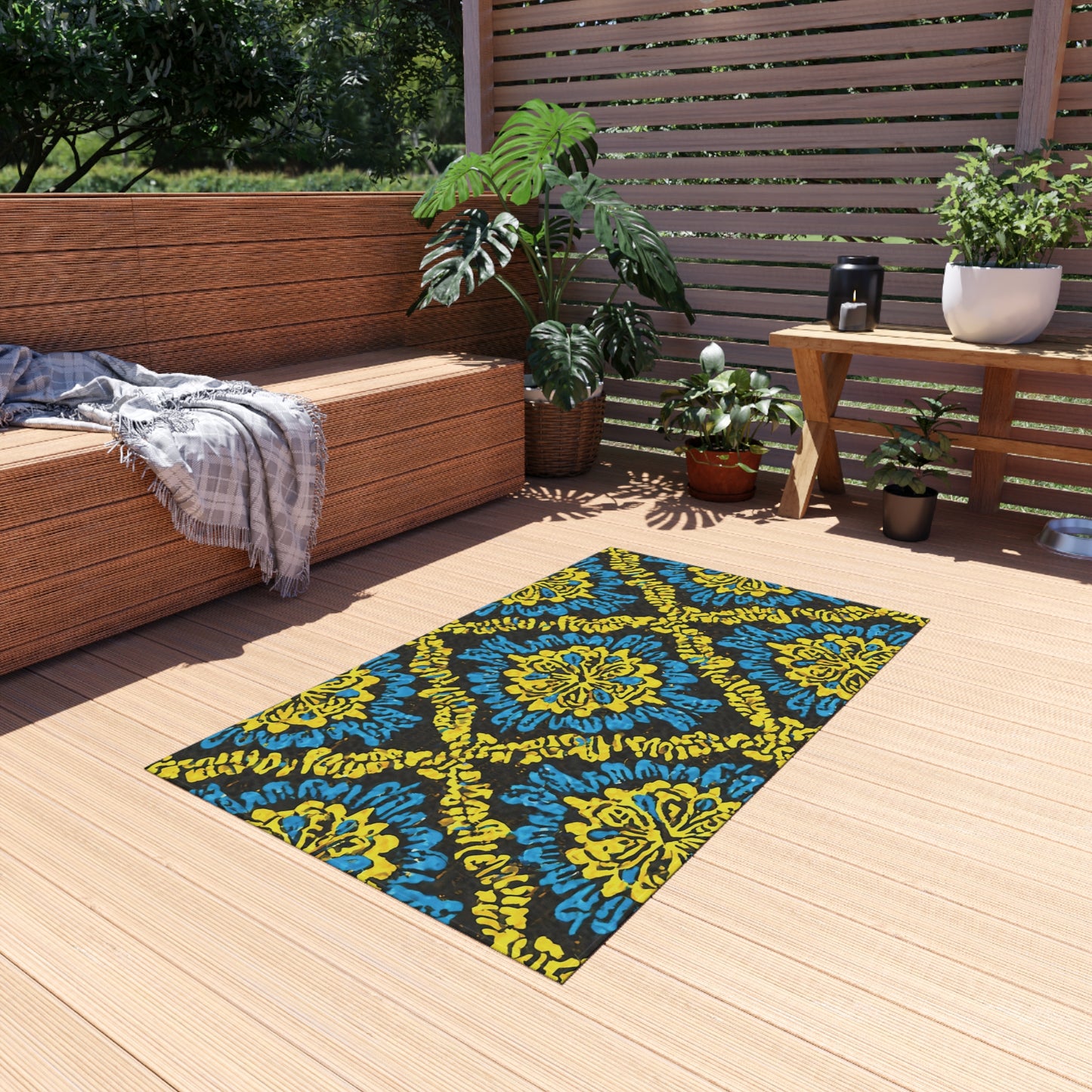 A weather-resistant patio rug with yellow and blue floral designs lies on a light wooden patio. Nearby are potted plants, a gray blanket draped over a bench, a lantern, and sunlight filtering through slatted privacy screens.