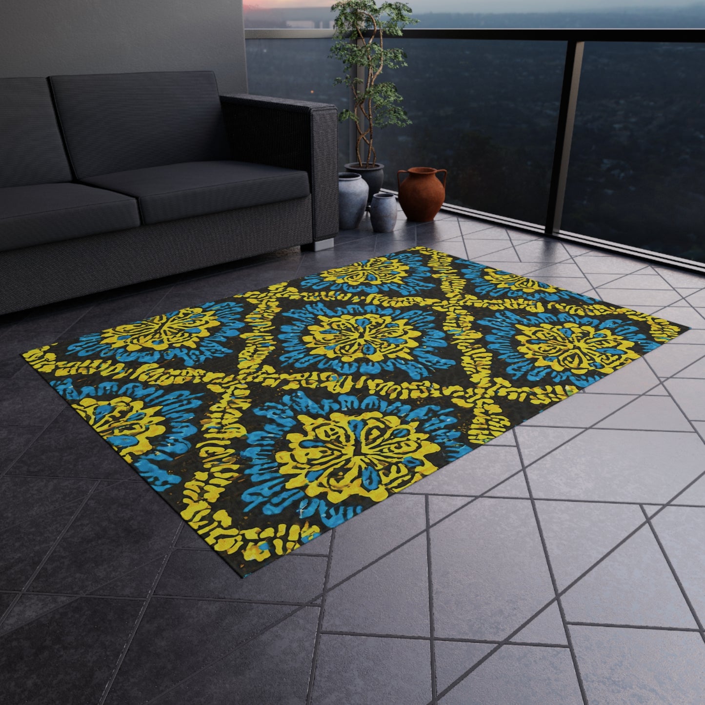 A modern balcony features a gray sectional sofa, an African-inspired outdoor rug with blue-and-yellow ornate patterns, two clay pots with leafy plants, and dark tiled flooring. A glass railing offers a cityscape view at dusk.