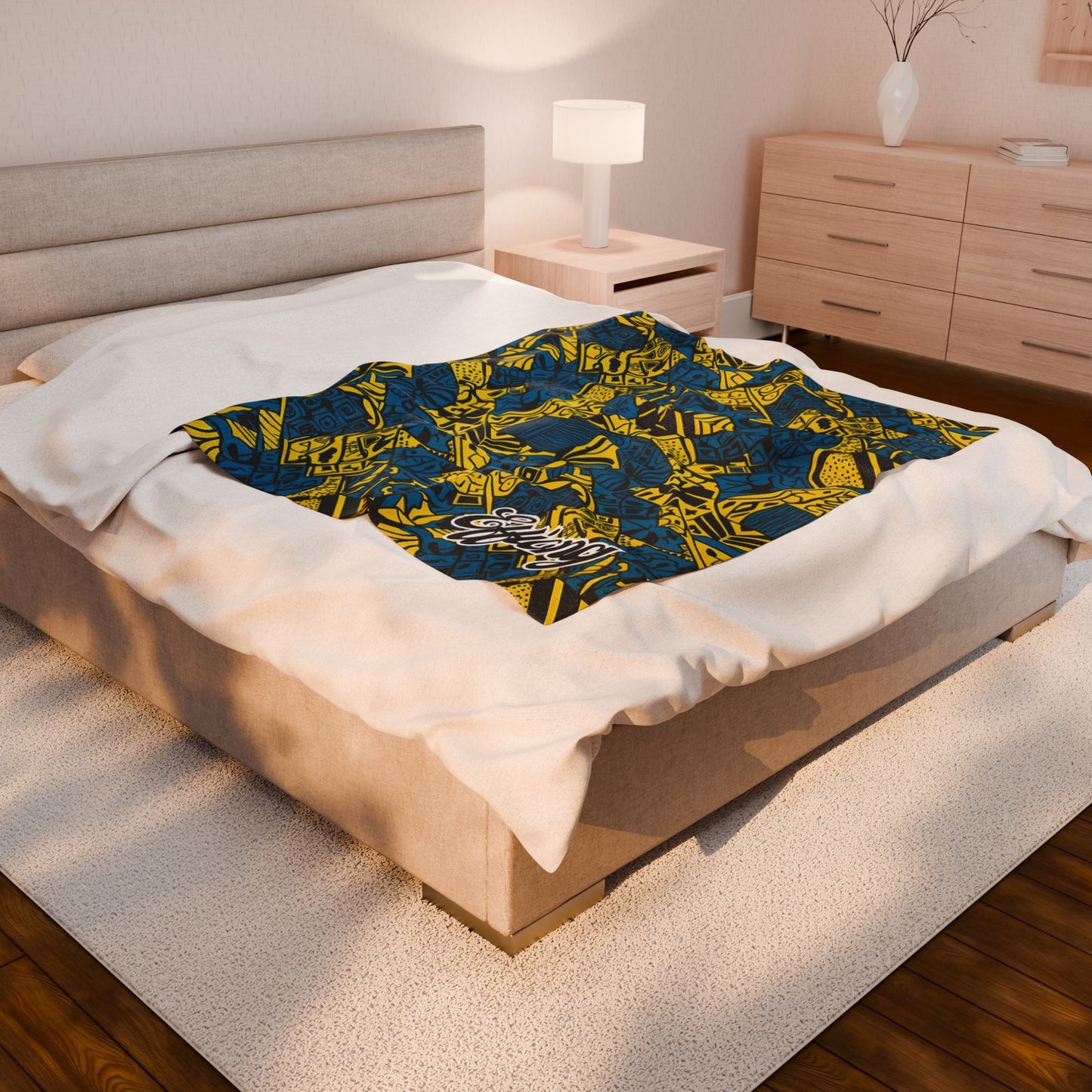 A modern bedroom features a beige upholstered bed with an African-inspired blanket in bold blue, yellow, and black patterns atop crisp white bedding. A wooden nightstand with a lamp, dresser, and white vase with branches complete the light, airy space.