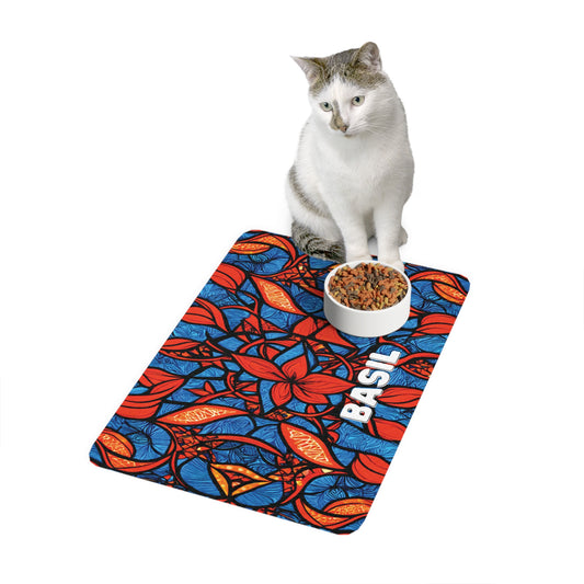 A white and gray cat sits behind a food bowl on a vibrant, African-inspired mat with red, blue, and orange abstract leaf patterns. The mat has the name BASIL printed in bold white letters. The background is plain white.