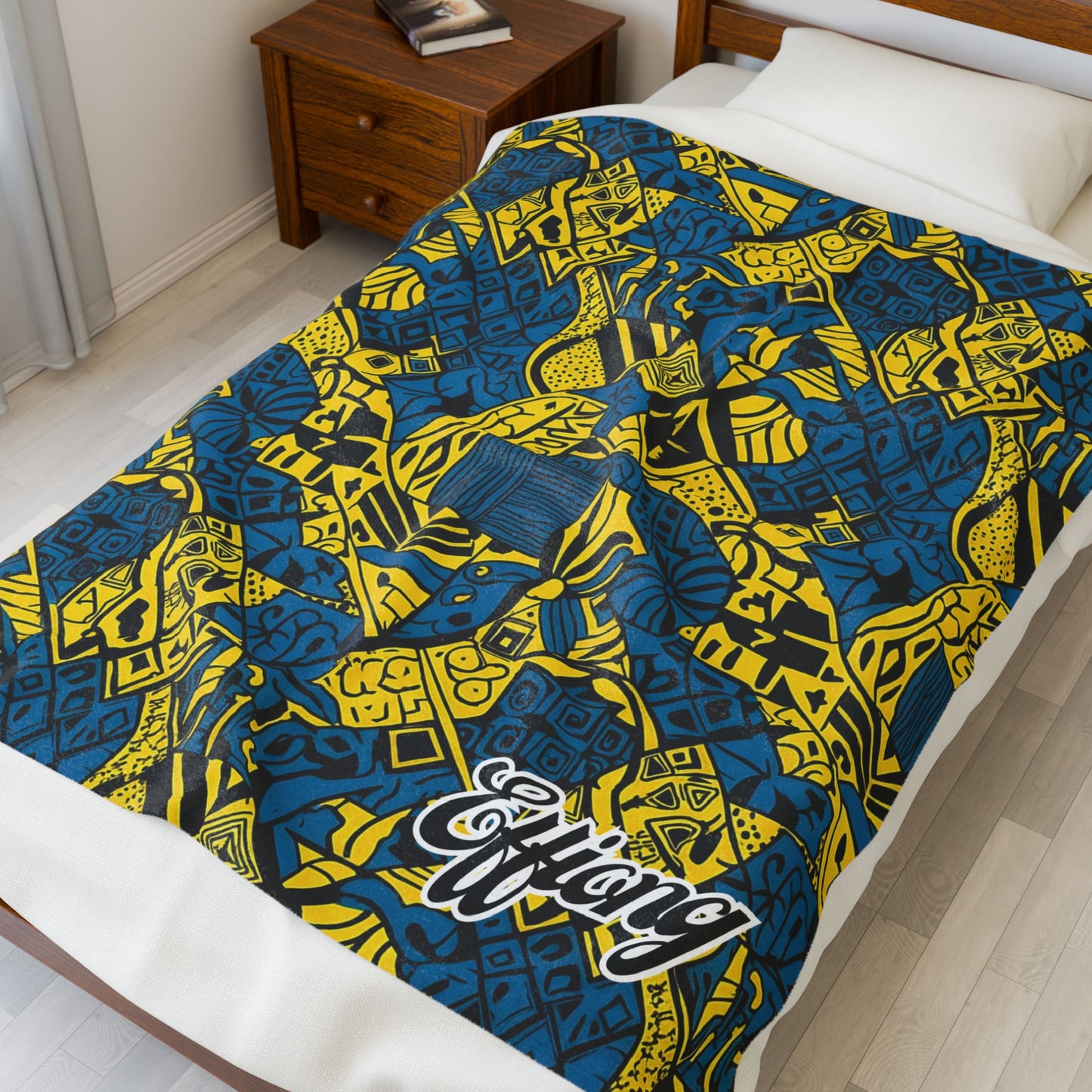 A bed with a colorful, African-inspired blanket featuring bold yellow and blue geometric patterns. This personalized velveteen blanket has Effiong in large, stylized white letters. The room includes light wood flooring and a wooden nightstand.