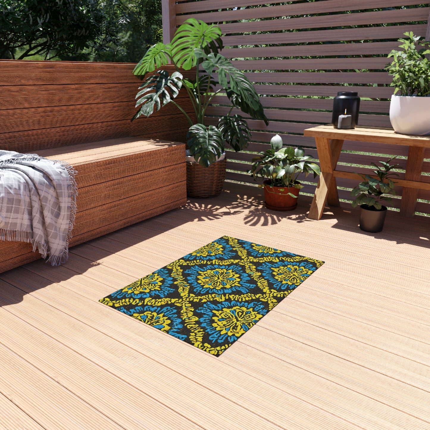 A small, colorful African-inspired outdoor rug with blue, yellow, and green patterns lies on a sunlit wooden deck. There’s a wooden bench with a gray plaid throw, potted plants, and a table with more plants and a black teapot. Slatted fencing provides partial shade.