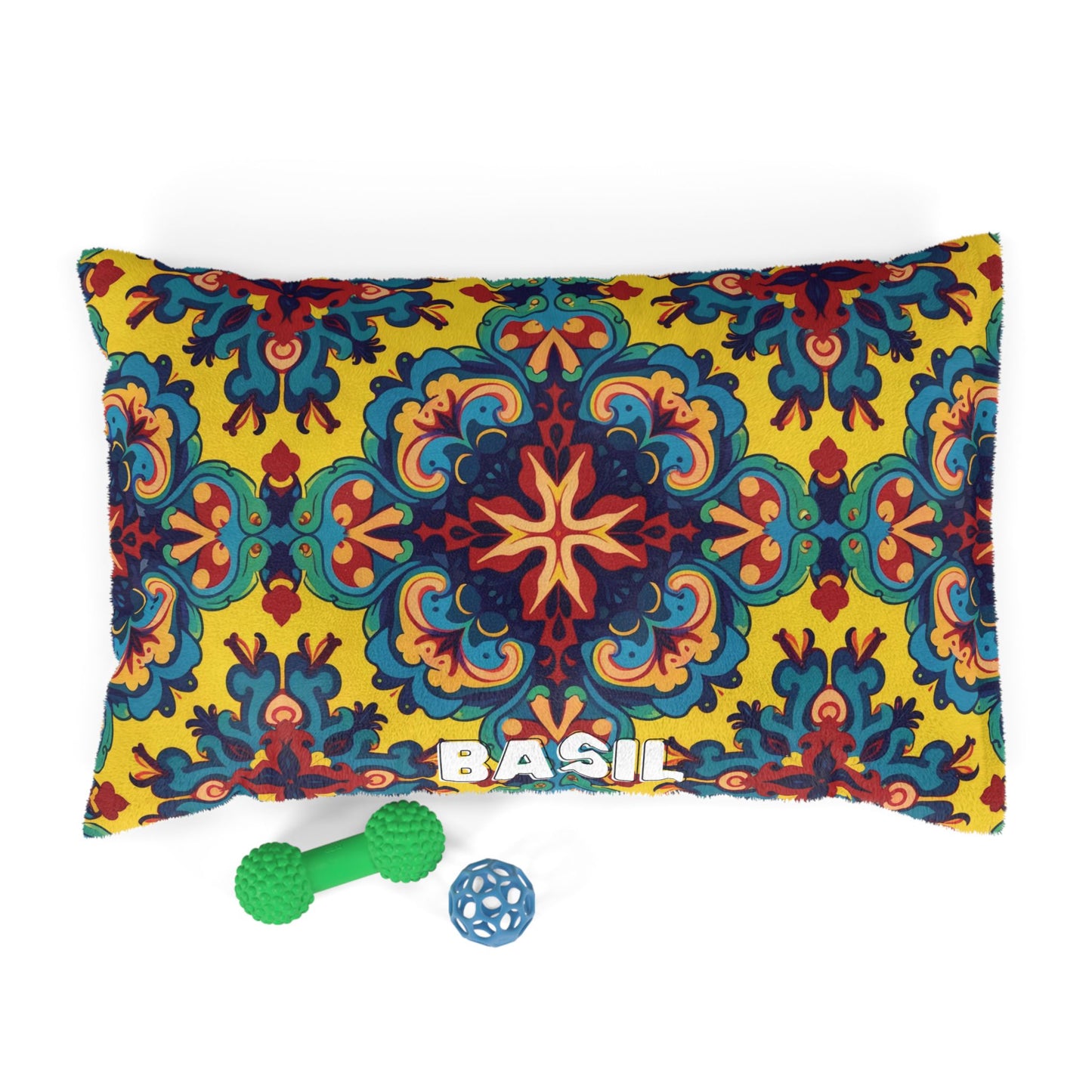 Rectangular custom dog pillow with a vibrant, multicolored pattern on a yellow background and the name BASIL in white letters. Below the personalized pet bed are two dog toys: a green dumbbell-shaped toy and a blue ball with cutout sections.