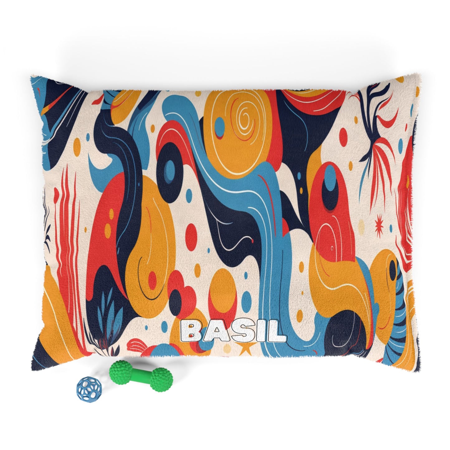 A rectangular pillow with “BASIL” in large white letters sits against a white background. The pillow features an African-inspired colorful abstract pattern with swirls and shapes in blue, yellow, red, and orange. Two small dog toys are nearby.