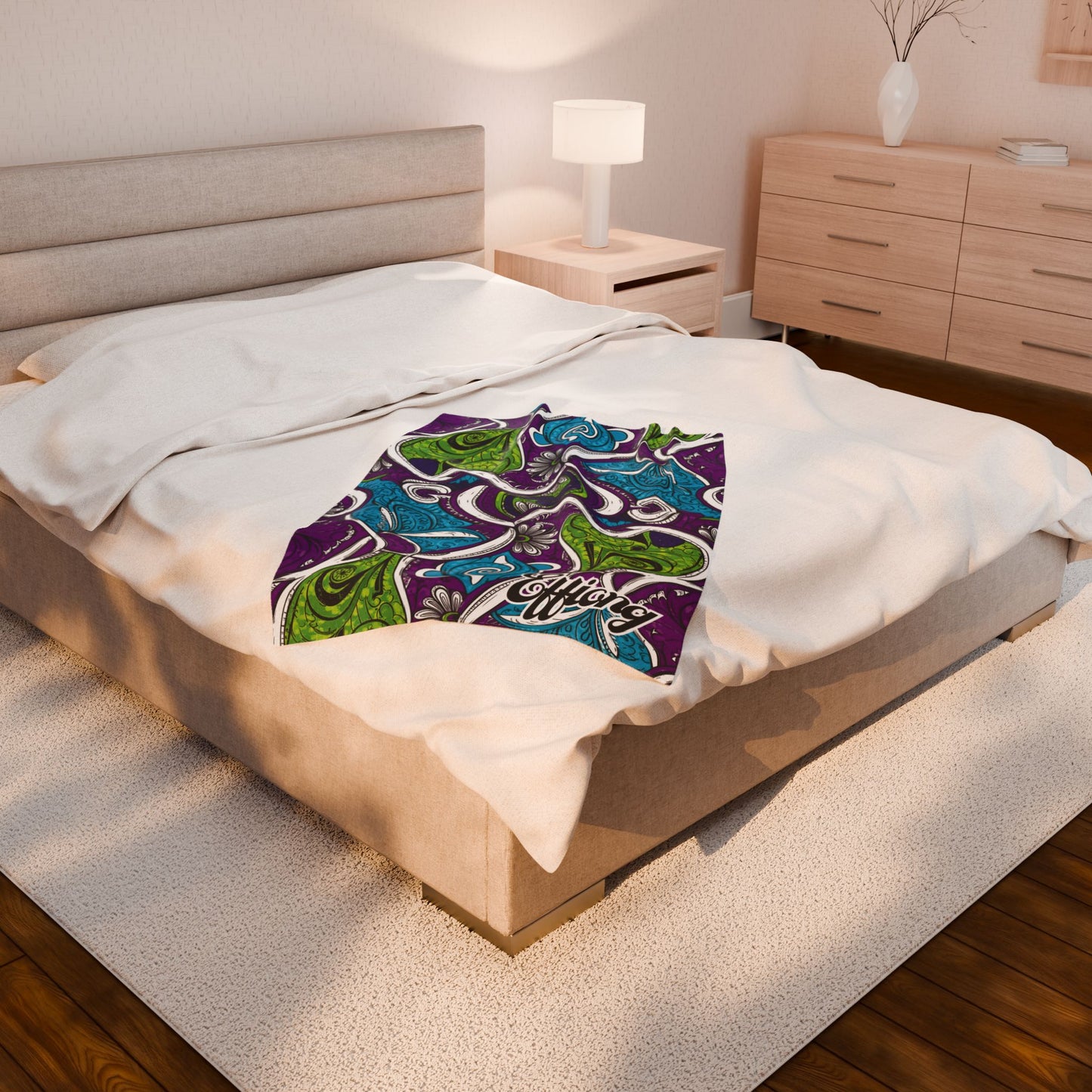 A beige bed with white bedding sits in a cozy, modern bedroom. An African-inspired blanket with green, blue, and purple swirls and “Good Vibes Only” lies on top. The room features wooden floors, a lamp, dresser, and potted plant.