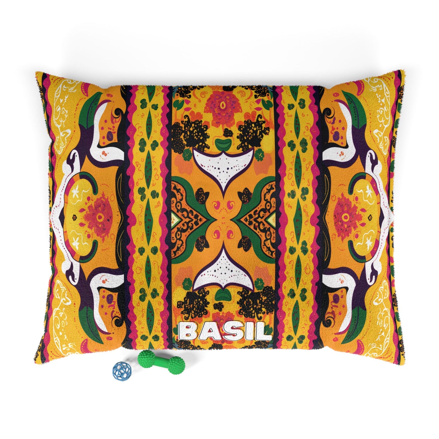 A colorful, African-inspired pet pillow with vibrant, symmetrical floral and geometric patterns in yellow, orange, green, and black. The pillow features “BASIL” in bold white letters. Two small pet toys—a green bone and a blue ball—are placed nearby.
