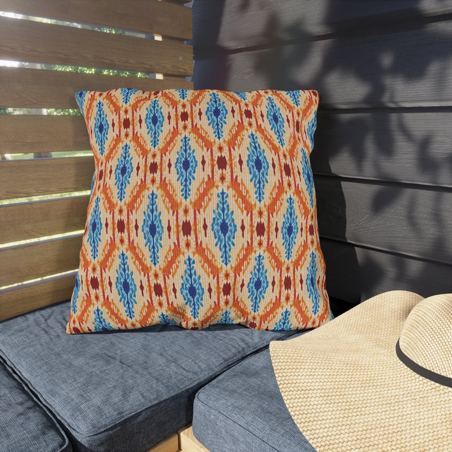A colorful, UV-resistant outdoor pillow with a geometric blue, orange, and red pattern rests on a blue bench next to a woven straw hat in a shaded patio area.