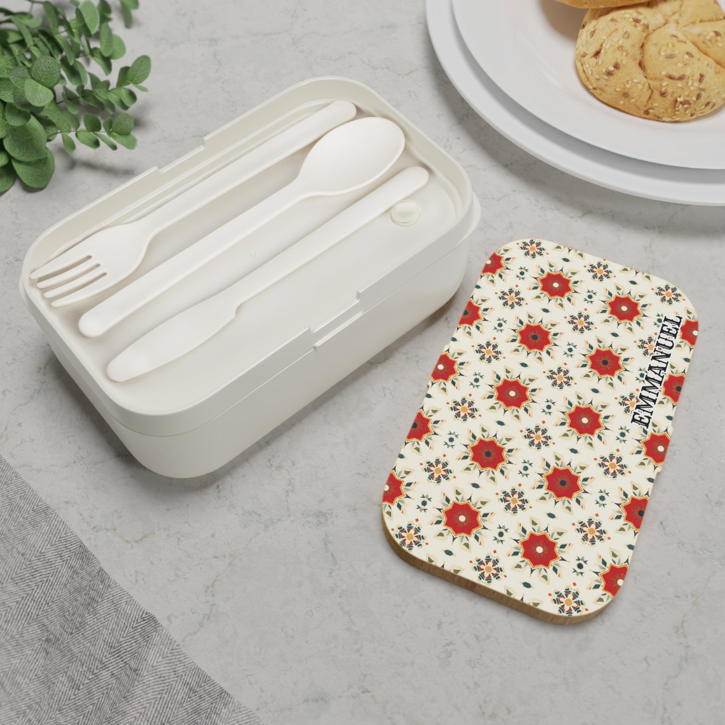 A white personalized bento box with a floral-patterned lid and the word EXEMPLARY sits on a light gray surface. Next to it, white plastic fork, spoon, and knife are neatly arranged with cookies and green leaves nearby.