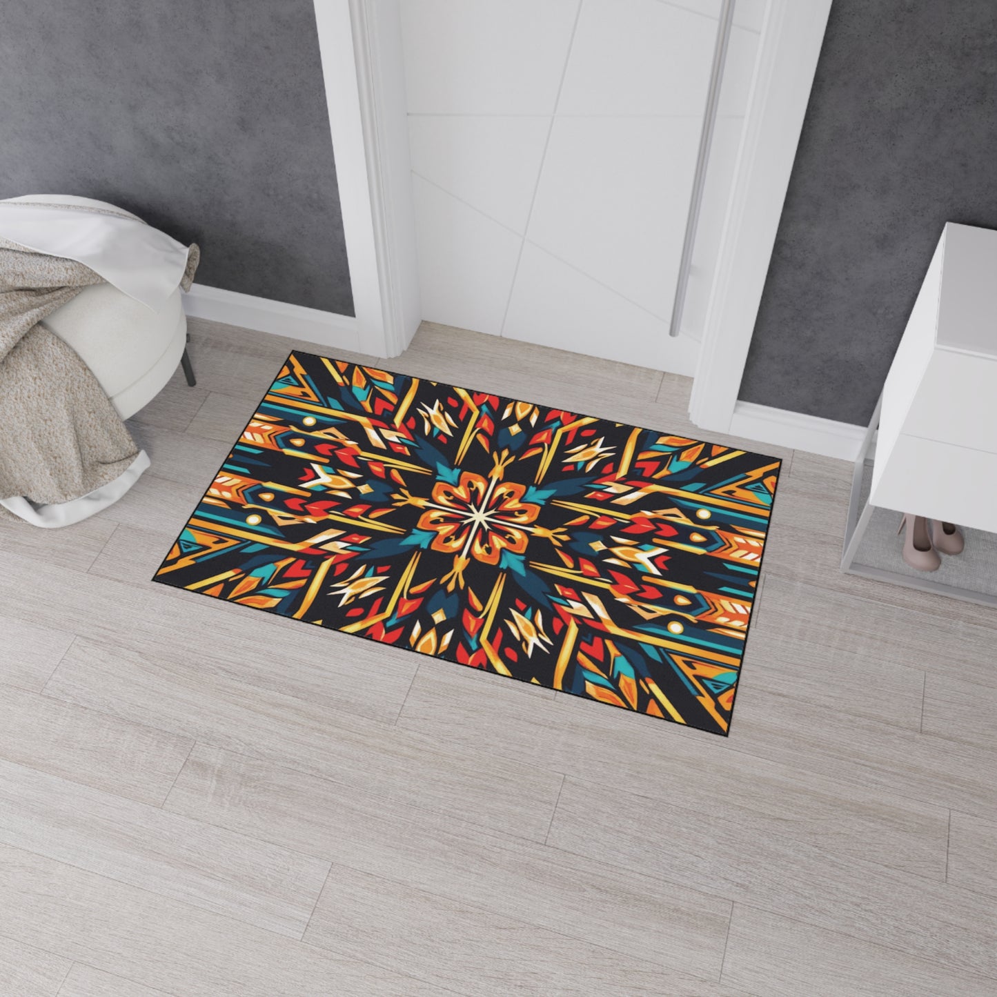 A colorful rectangular rug with bold geometric and floral mandala patterns in orange, yellow, blue, red, and black lies on a light wood floor—its non-slip rubber backing keeps it secure next to a folded beige blanket and small white cabinet with shoes.