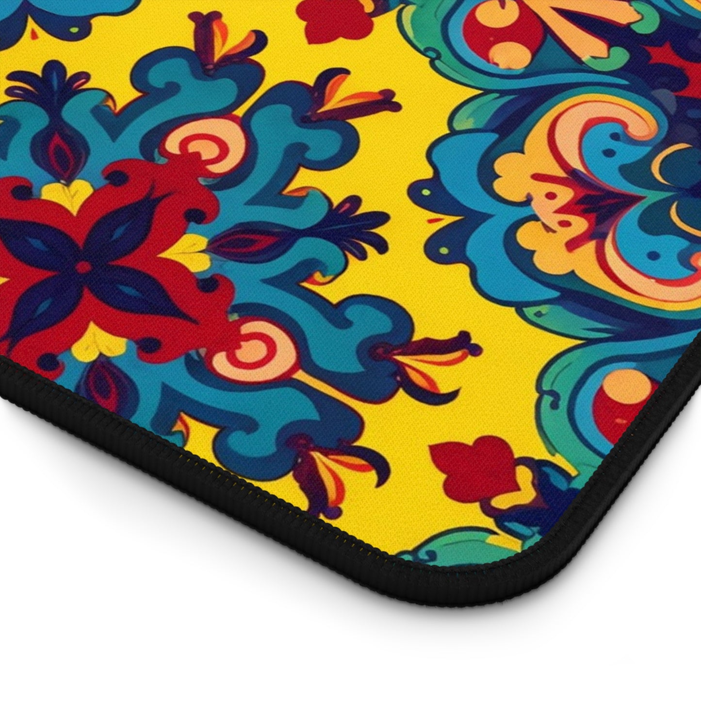 A close-up of a neoprene desk mat corner showcases vibrant, ornate floral patterns in red, blue, and teal on a bright yellow background. The design features black trim and decorative swirls—perfect as a colorful workspace accessory.