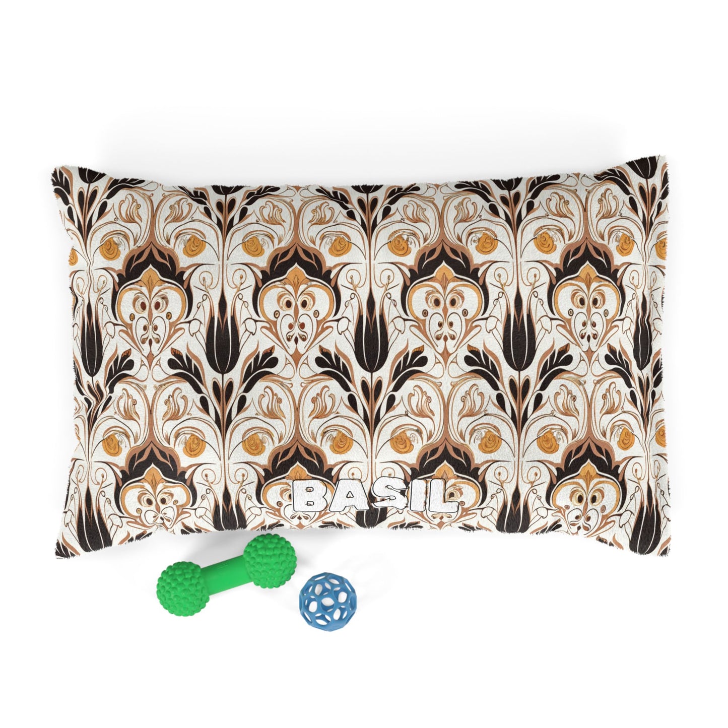 A rectangular pillow featuring an African-inspired beige, black, and gold ornate floral pattern displays the name BASIL in bold white letters. In front are two green textured dog toys and a blue ball with cutout designs, set against a white background.