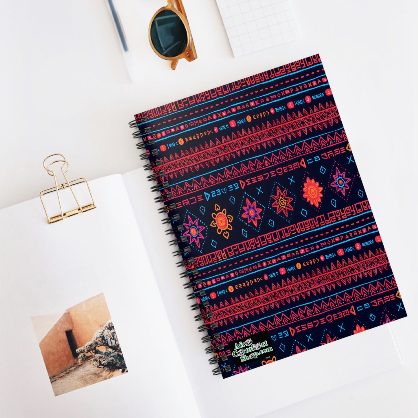 A spiral notebook with a dark cover featuring bright, culturally inspired geometric and floral patterns lies on a white desk. Nearby are an open book displaying a photo, gold binder clip, sunglasses, and a small grid notepad.