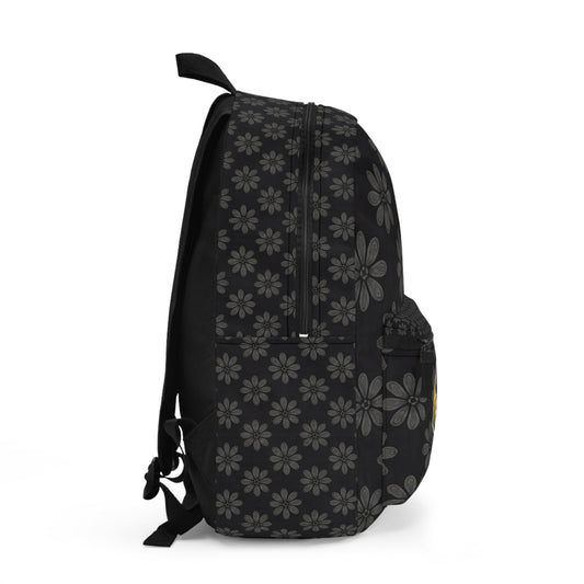 A black boho chic rucksack viewed from the side, featuring a repeating pattern of gray daisy-like flowers. The bag has a front pocket, mesh side pocket, zippers on top, and adjustable shoulder straps visible on the left. Plain white background.
